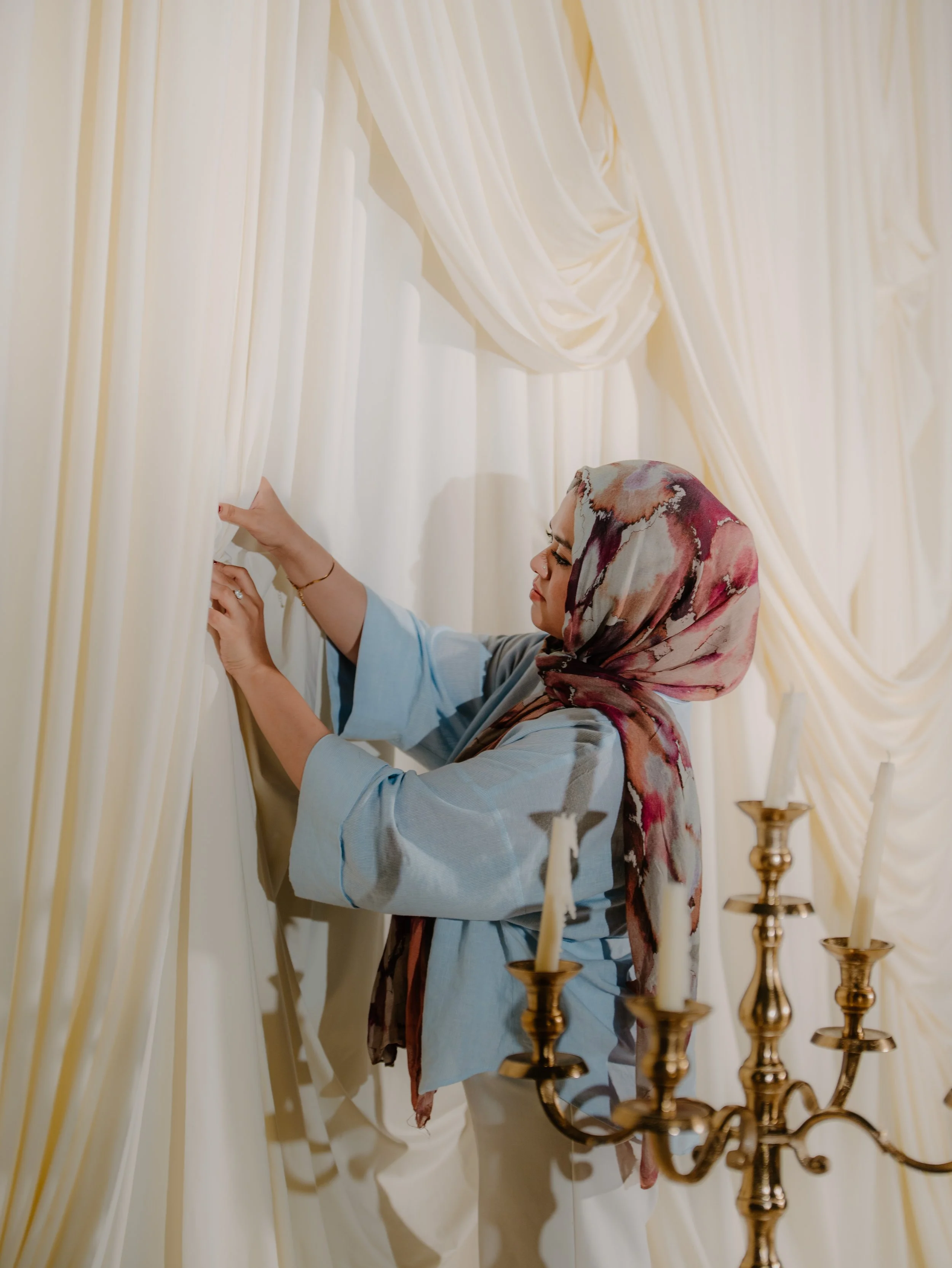 A woman in a light blue outfit and a colorful headscarf adjusts cream-colored curtains behind a chandelier with candles.