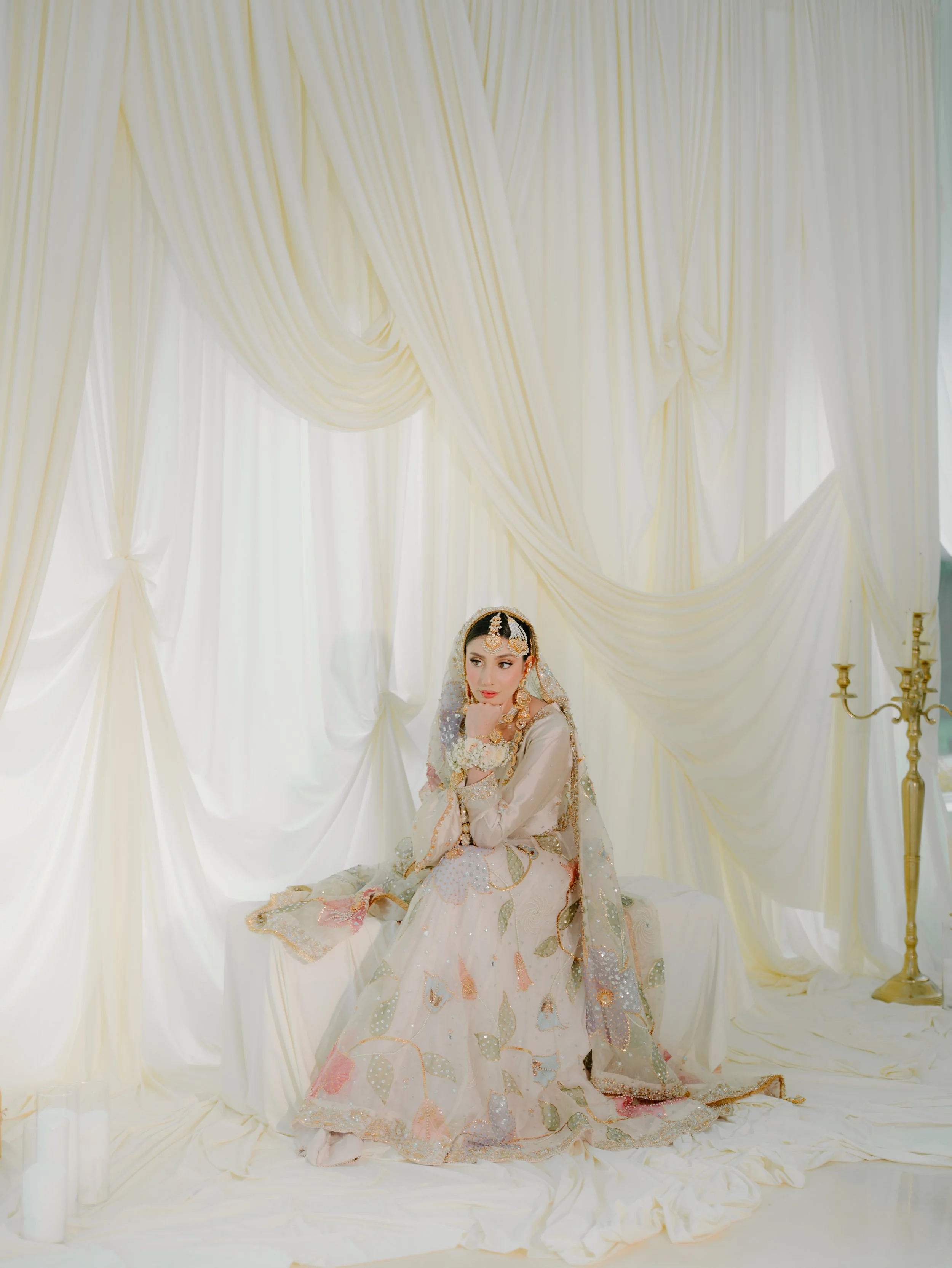 A bride in traditional attire sitting on a white cloth-covered surface in front of cream-colored draped curtains with a candelabrum to her right, posing indoors.