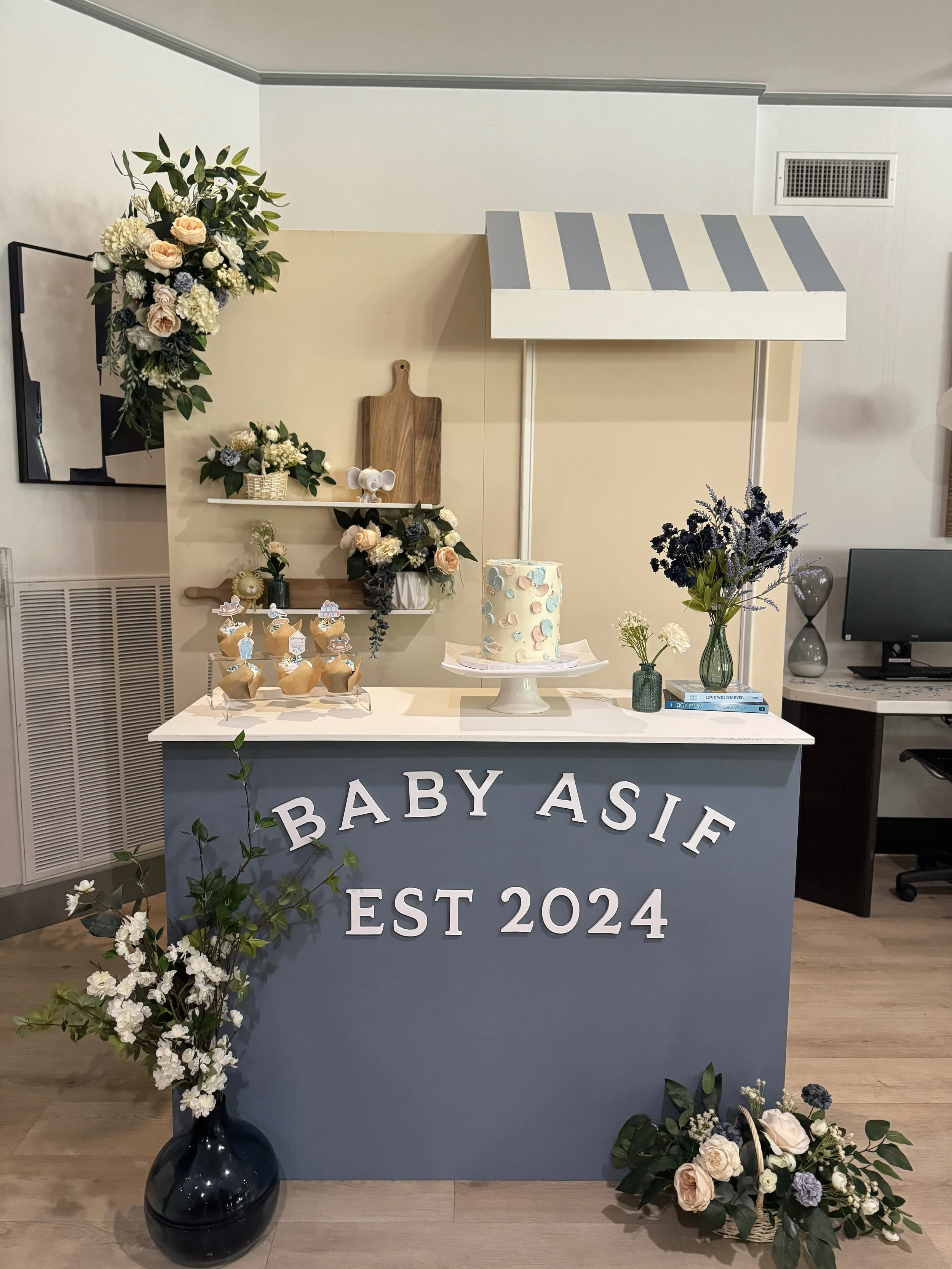 A baby shower setup with a blue and white theme, featuring a cake, cupcakes, floral arrangements, and a sign reading 'Baby Asif EST 2024' on the front of a table.