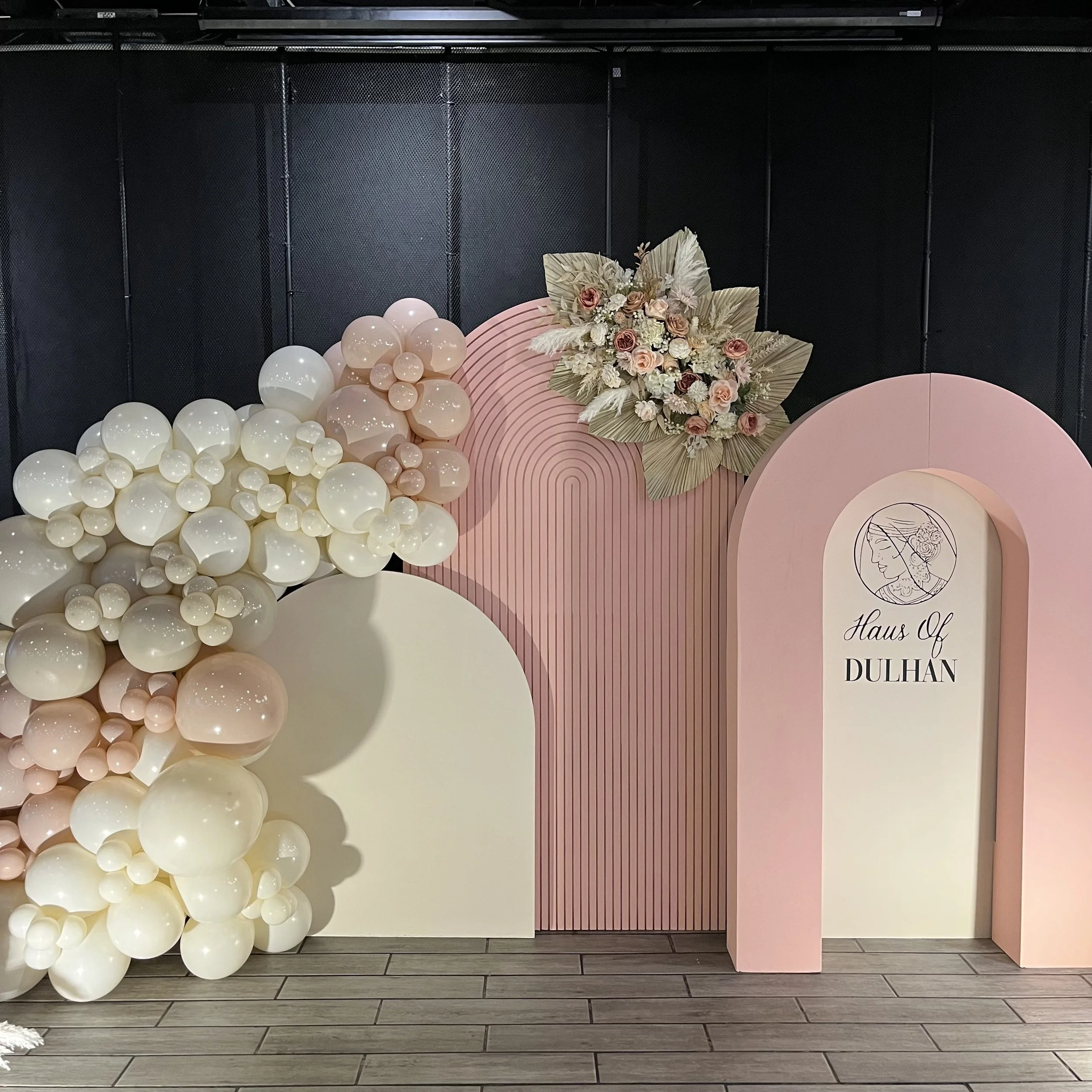 Decorative backdrop with balloons and floral arrangement for Haus of Dulhan event, featuring pink, white, and cream colors.