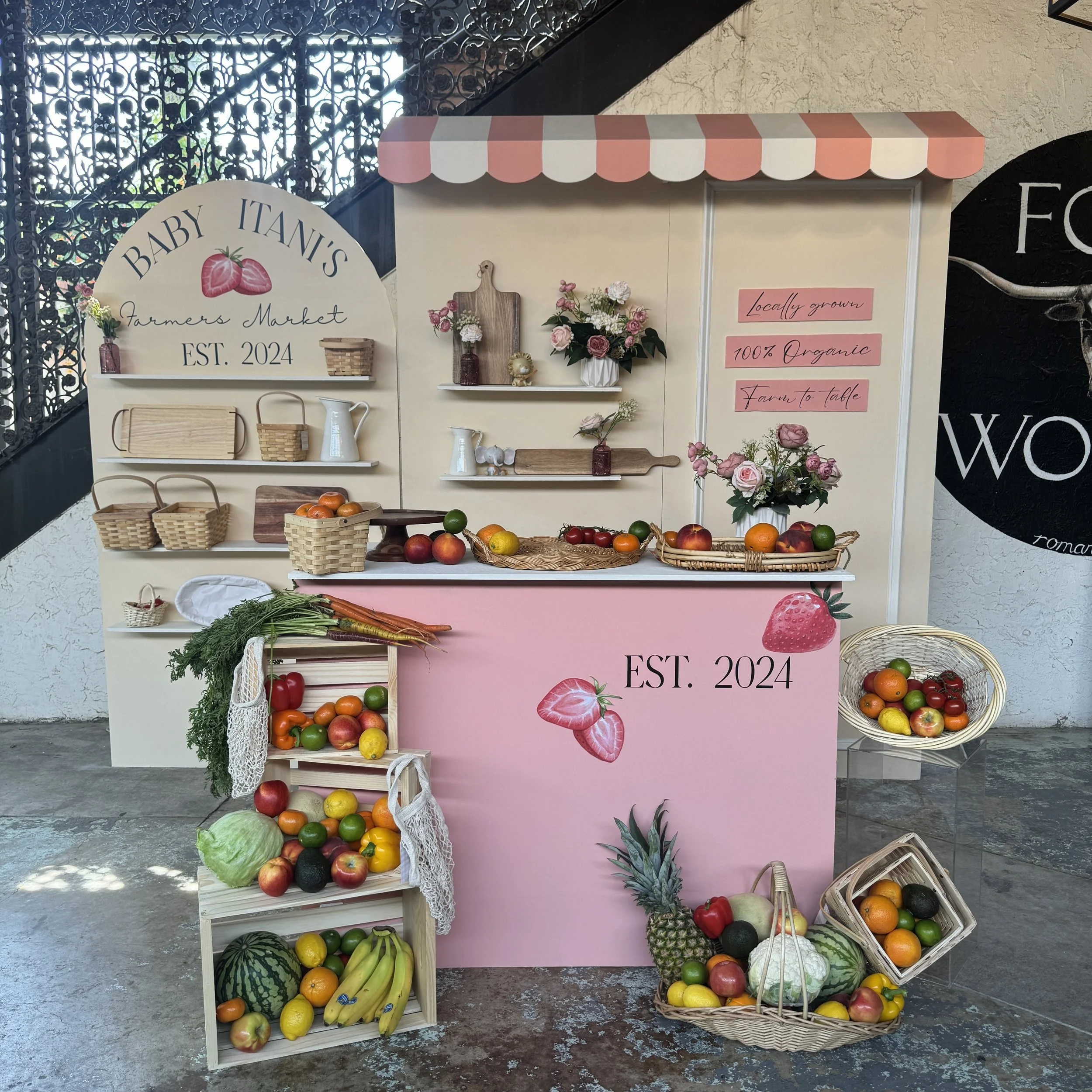 An outdoor farmer's market stand selling a variety of fresh fruits and vegetables, decorated with flowers and signs indicating it is locally grown, organic, and starting in 2024.