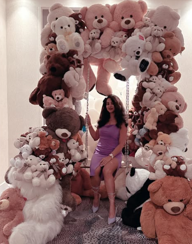 A woman in a purple dress sitting on a swing surrounded by a large collection of teddy bears and stuffed animals forming a bear-shaped frame.