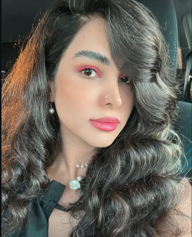 Close-up portrait of a young woman with long, wavy dark hair, wearing pink eye makeup, red lipstick, pearl earrings, a pearl necklace, and a small nose ring.