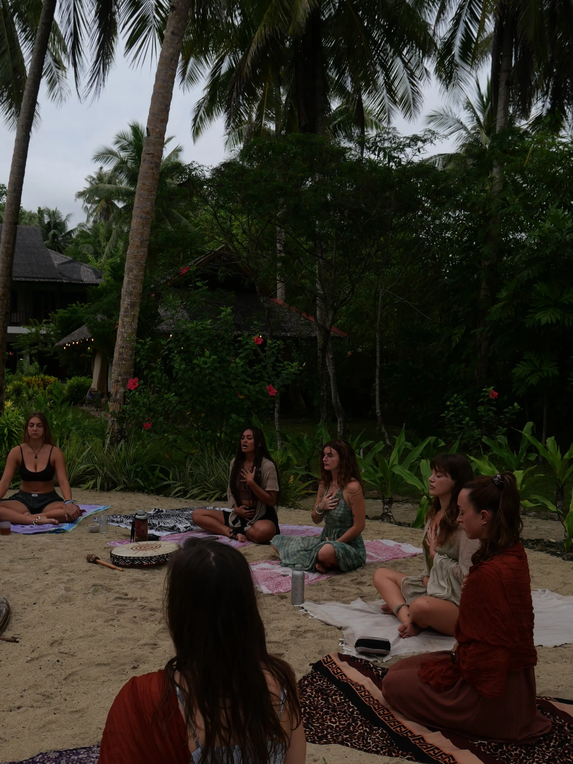 Group of women being led by Kaylani during a retreat in Siargao Island, practicing meditation on a beach with lush green trees and tropical plants in the background.