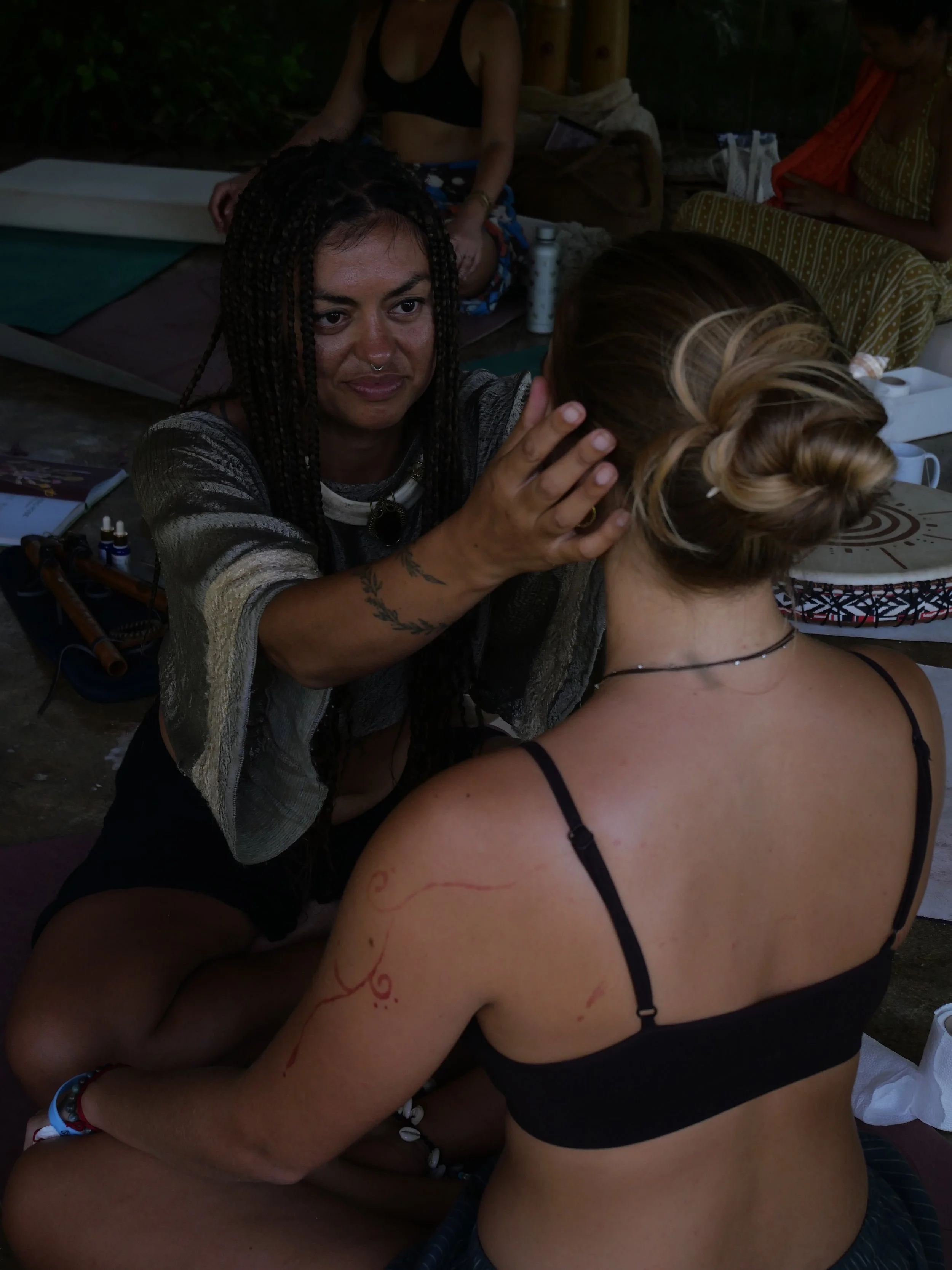 A woman with dark braided hair performs a spiritual or healing ritual on another woman with blonde hair styled in a messy bun. They are seated outdoors with other people seated nearby, surrounded by drums, bottles, and personal items, suggesting a gr