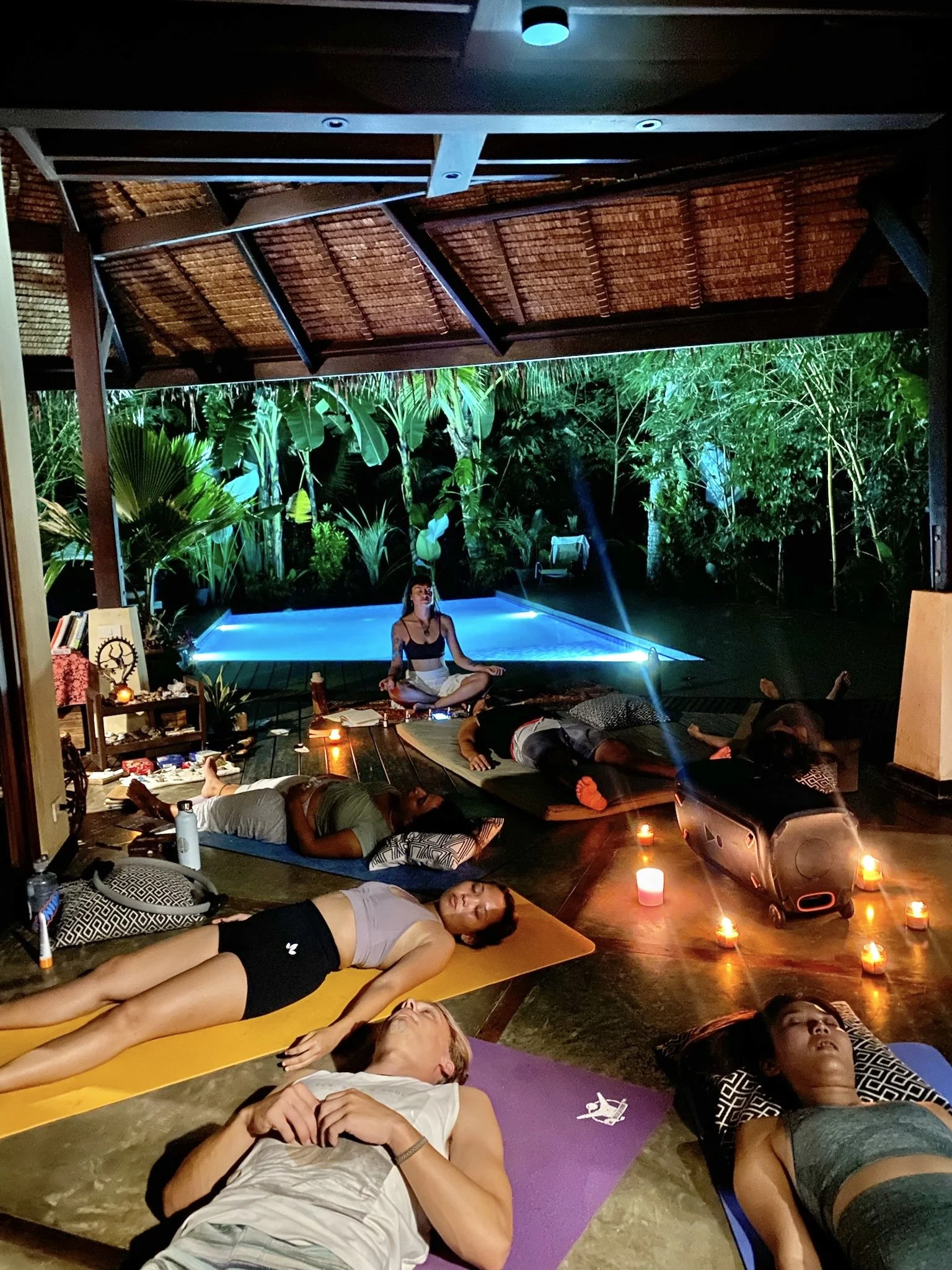 People participate in a nighttime outdoor yoga or meditation session on mats, candles, and a small pool in a lush, tropical setting with greenery.