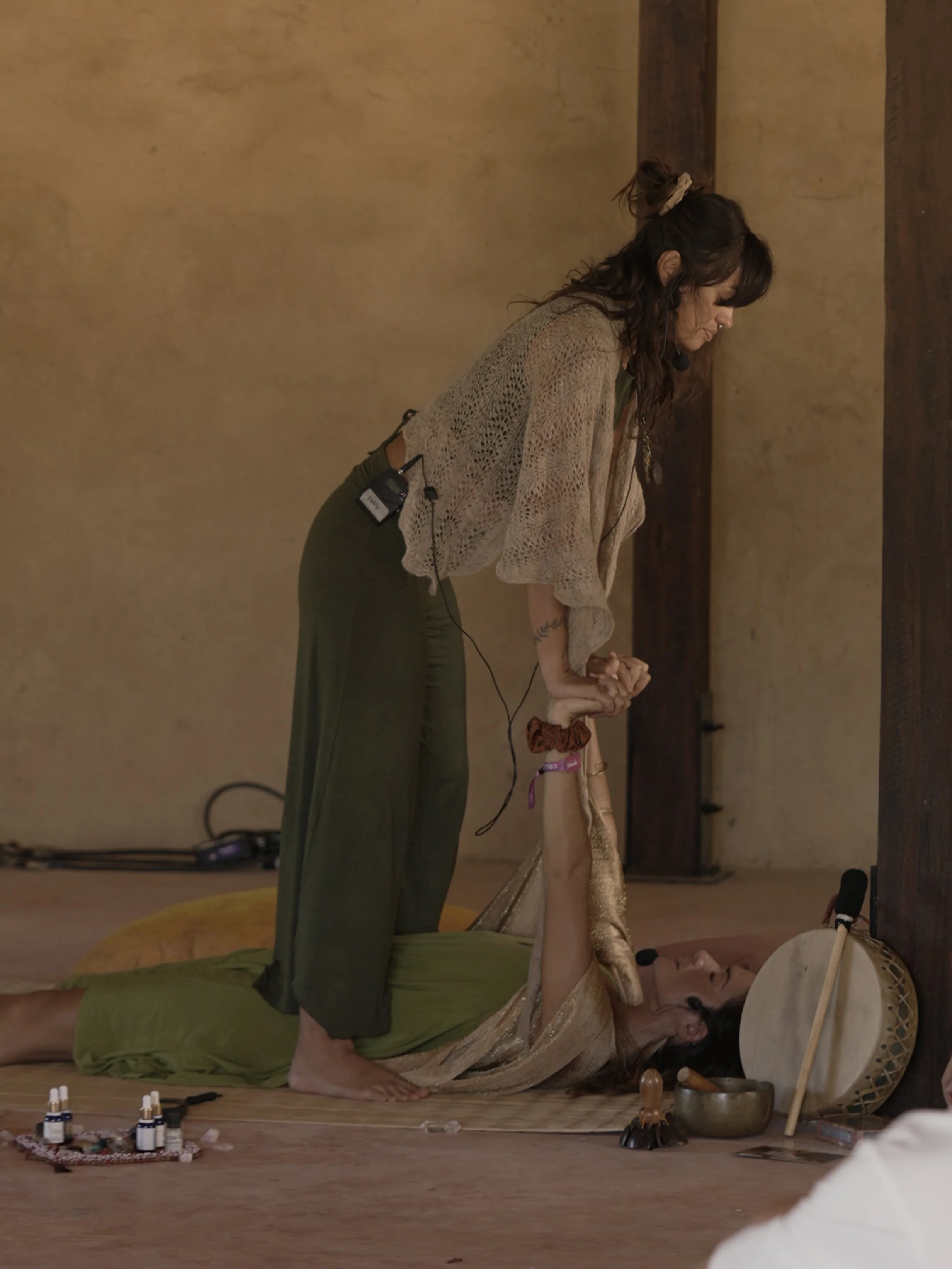 A woman is lying on the floor with a drum in front of her face, eyes closed, and another woman is standing and leaning over her, holding her hands. Taken during Wonderfruit Festival in a breathwork ceremony led by Kaylani Rhythms and Tamara.