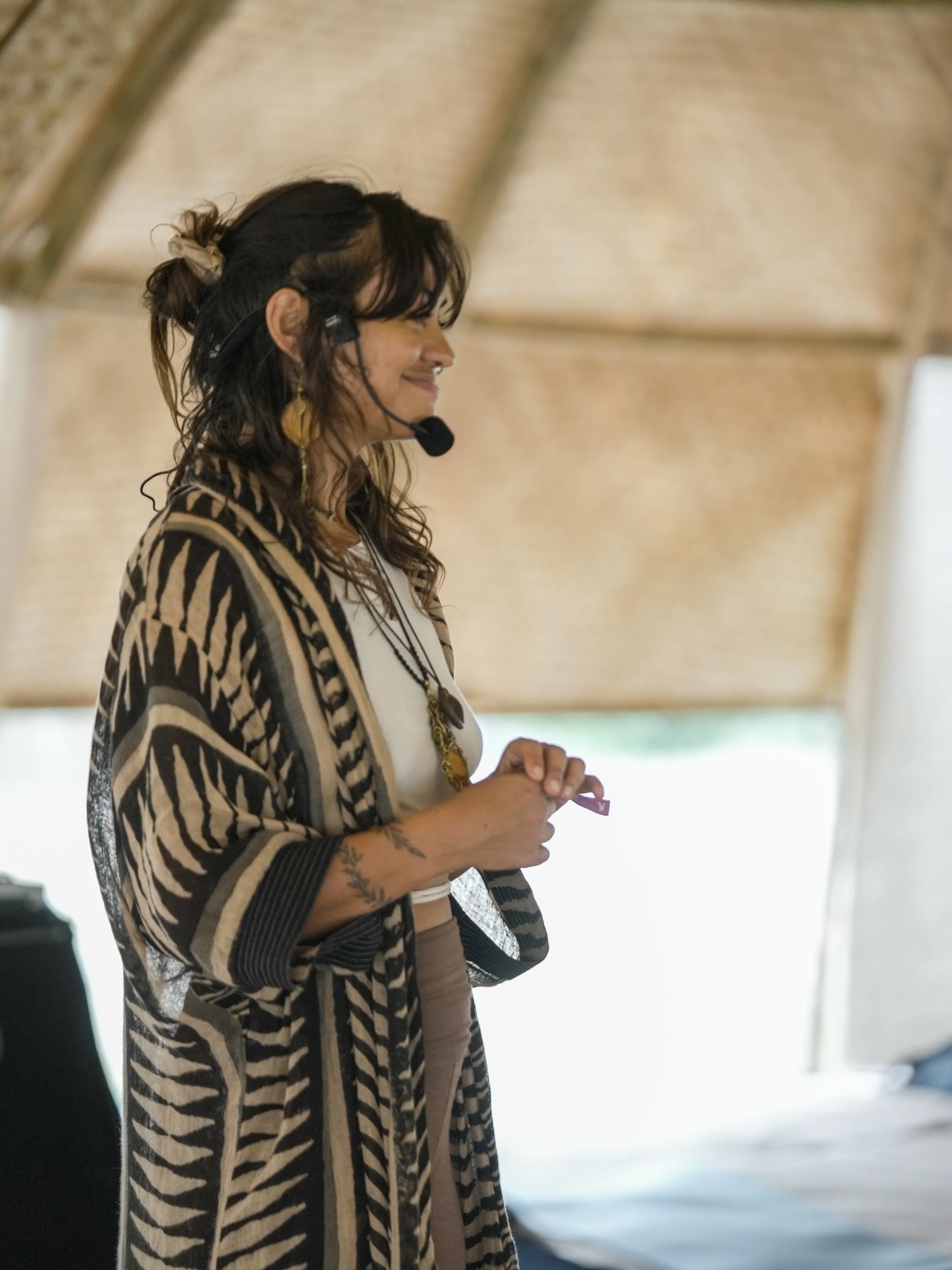A woman wearing a patterned kimono-style robe, standing inside a wooden pavilion, smiling, with a headset on, holding her hands together.