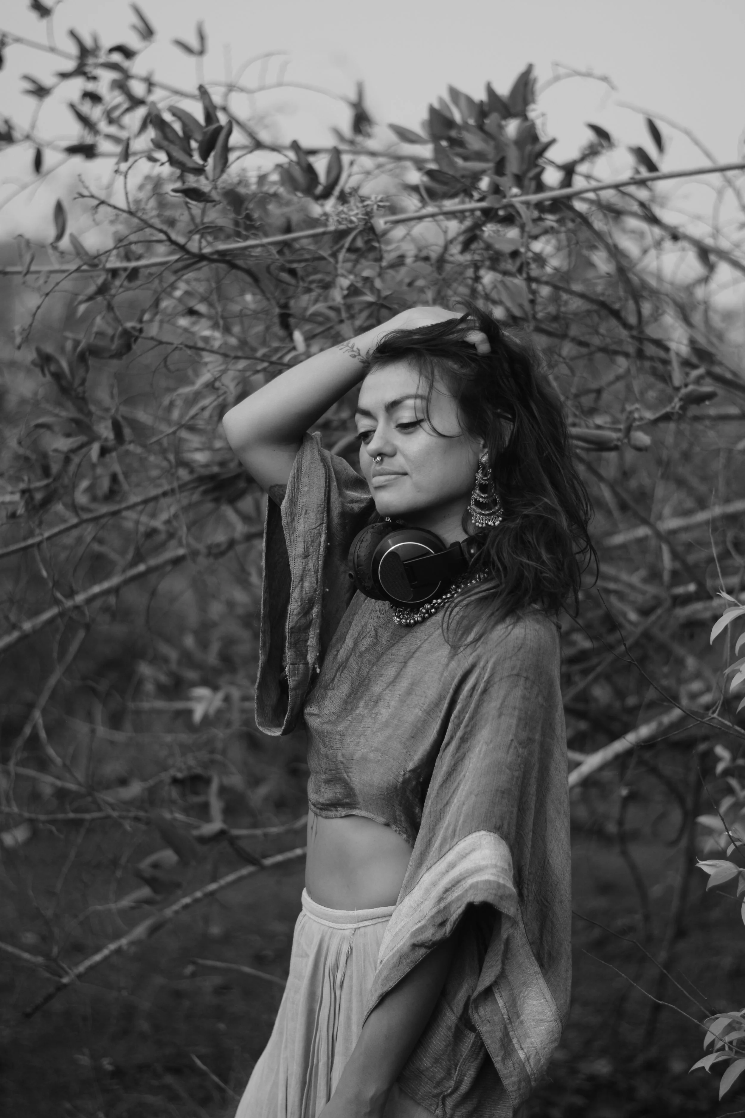 Kaylani standing outdoors among tree branches, with one hand in her hair, wearing a crop top, earrings, a necklace, and headphones around her neck, captured in black and white.