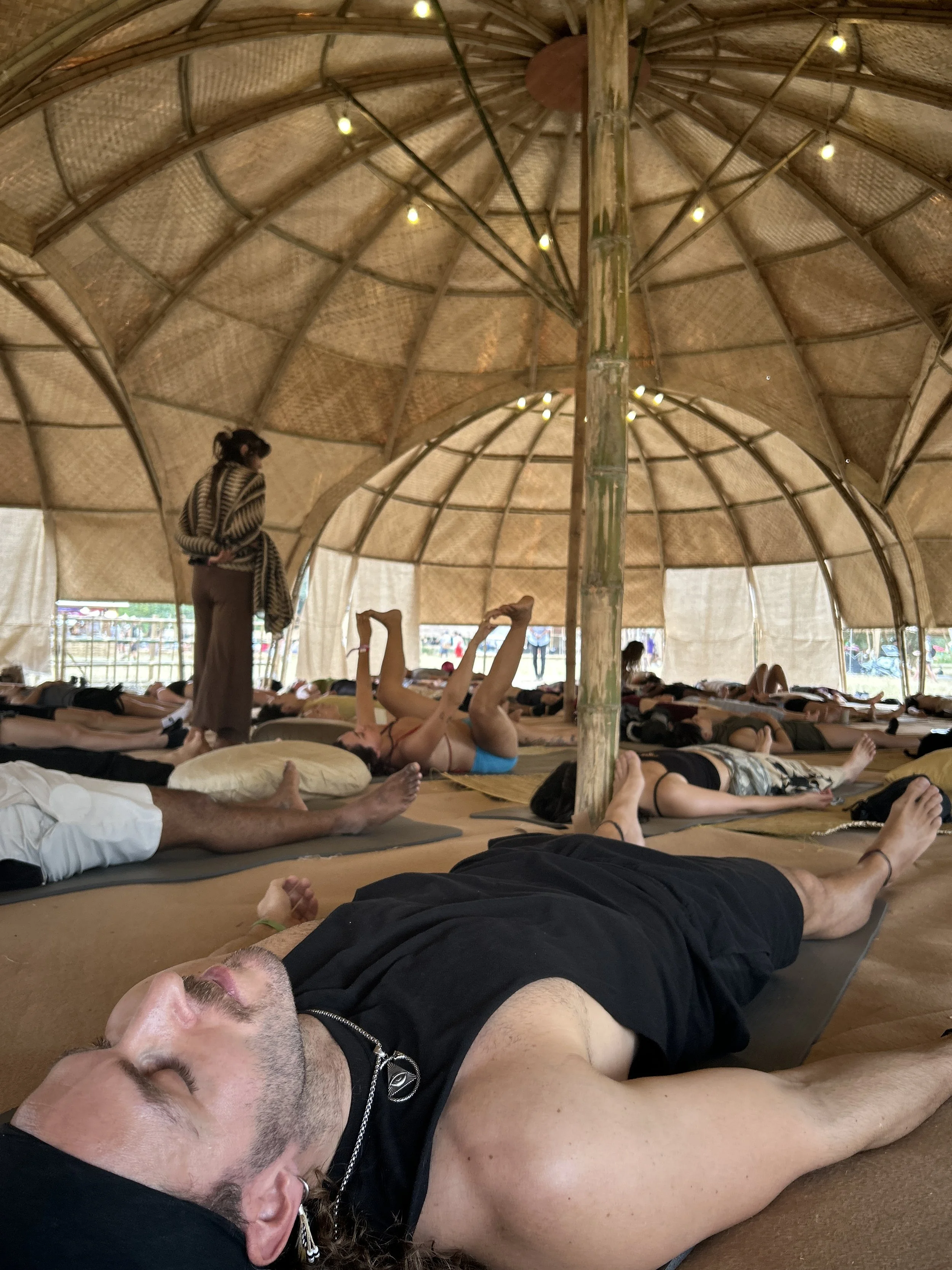 People practicing yoga or meditation inside a bamboo structure with a woven roof and string lights.