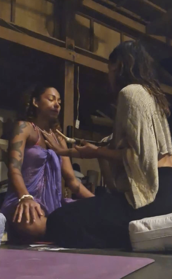 Two women in a dimly lit room, one wearing a purple dress with tattoos, sitting cross-legged, receiving a spiritual or healing treatment from the other woman in a gold top and black pants.