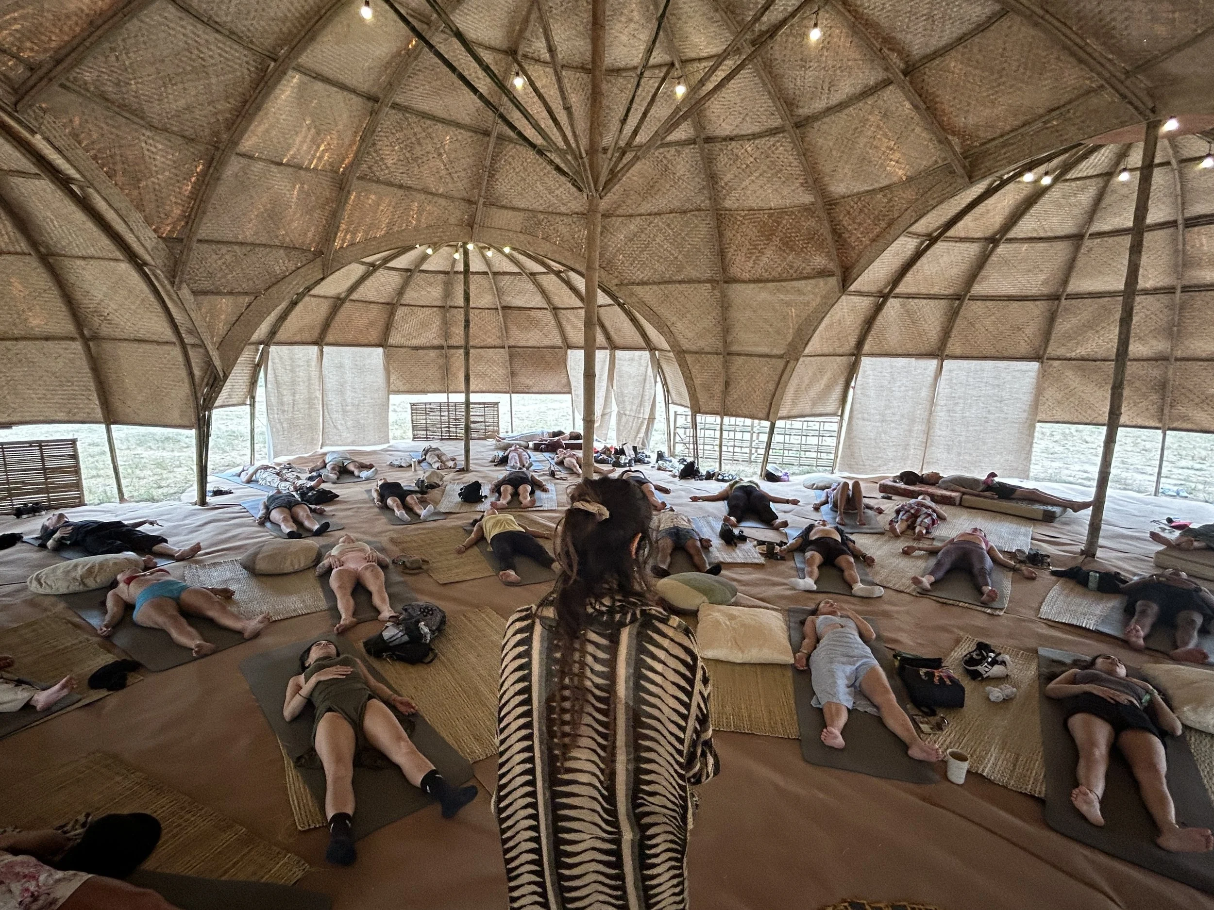 A group of people lying on mats and pillows inside a large, open-sided, thatched-roof structure, possibly participating in a yoga or meditation session.
