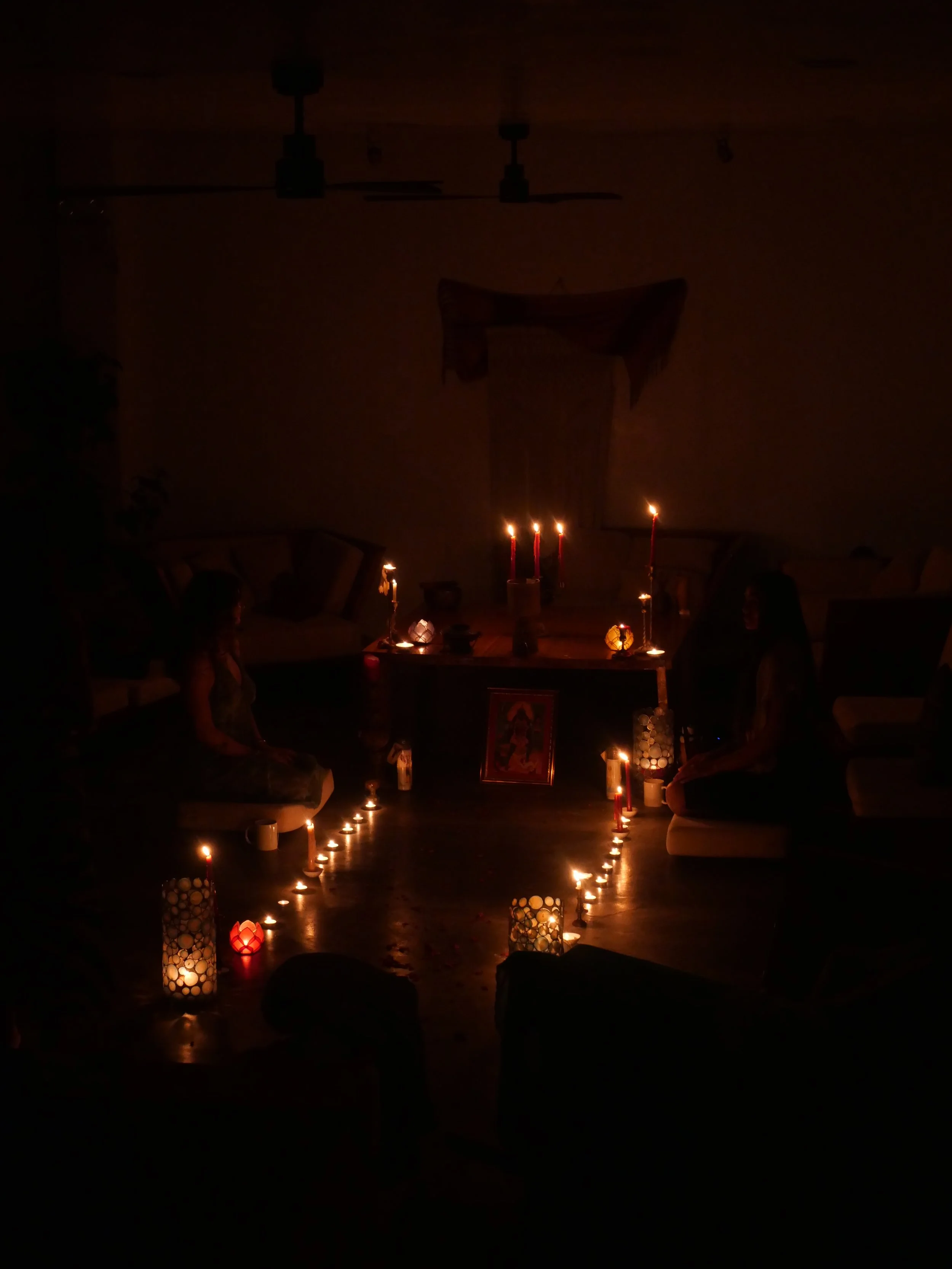 Dimly lit room with lit candles arranged in a heart shape on the floor, two people sitting on either side, and a small altar or picture on the floor in the center, during a retreat meditation session led by Kaylani Rhythms.
