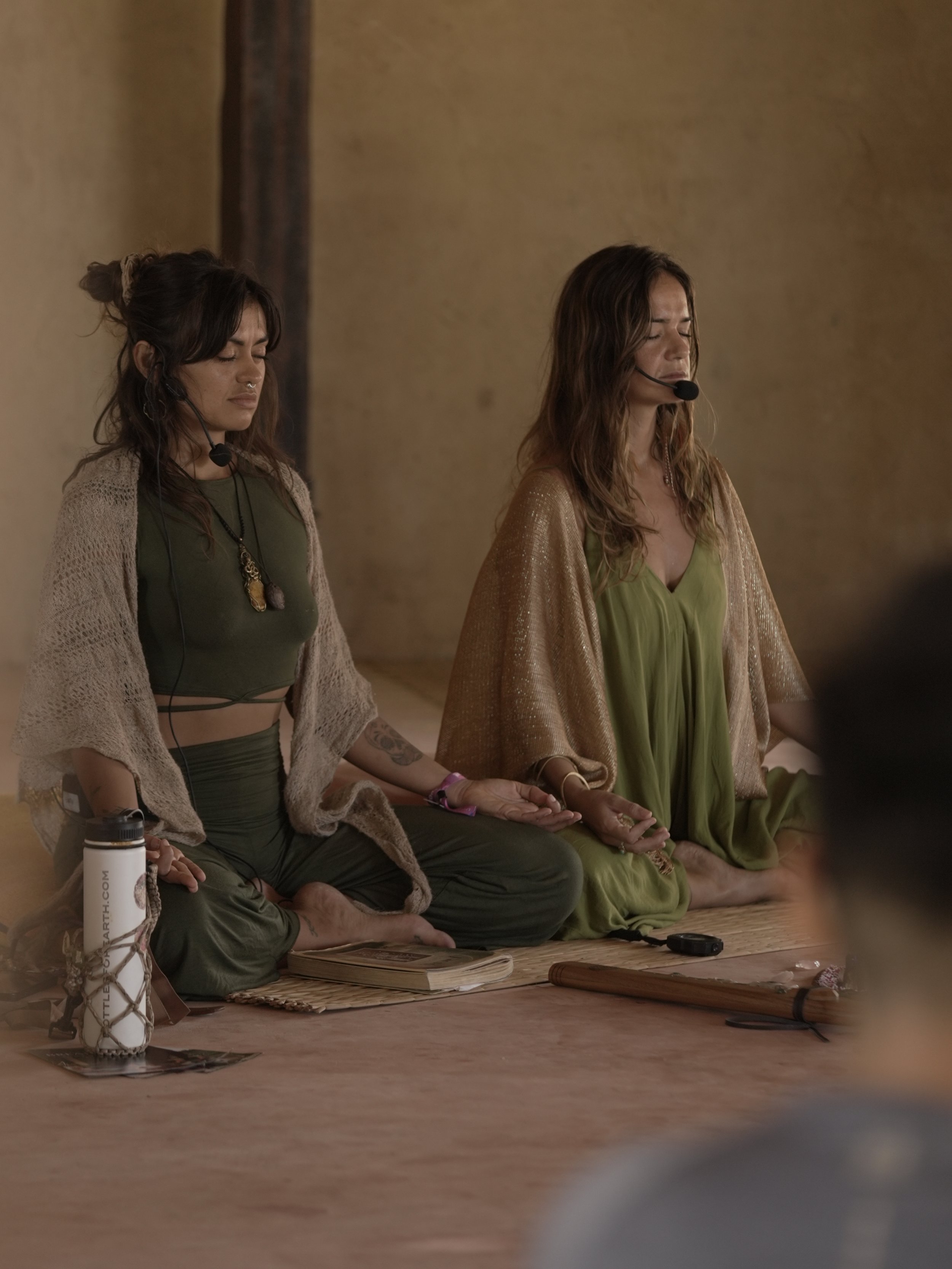Two women sitting cross-legged on the floor in prayer or meditation, eyes closed, with meditation accessories around them, in a room with plain walls.