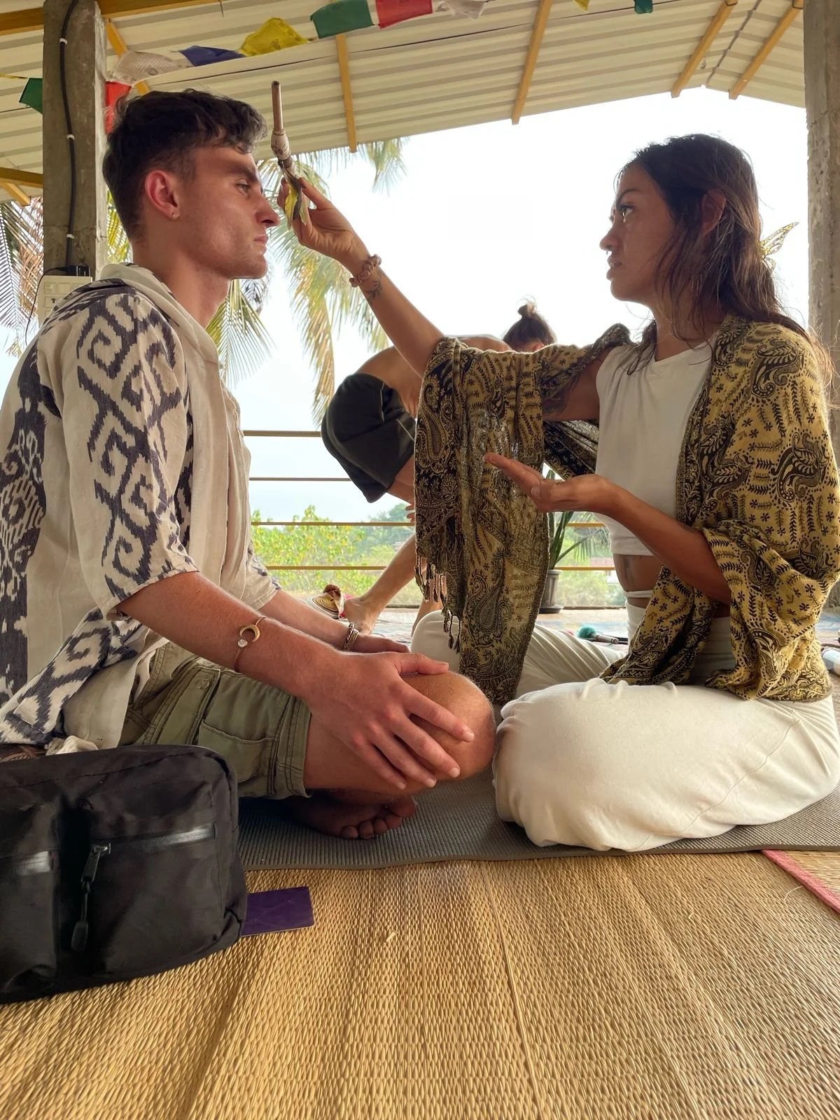 A woman with long hair, wearing a patterned shawl and white pants, is performing a ritual or ceremony on a seated young man. The man has short hair, is wearing a beige jacket and shorts, and is sitting cross-legged. The setting appears to be open-air