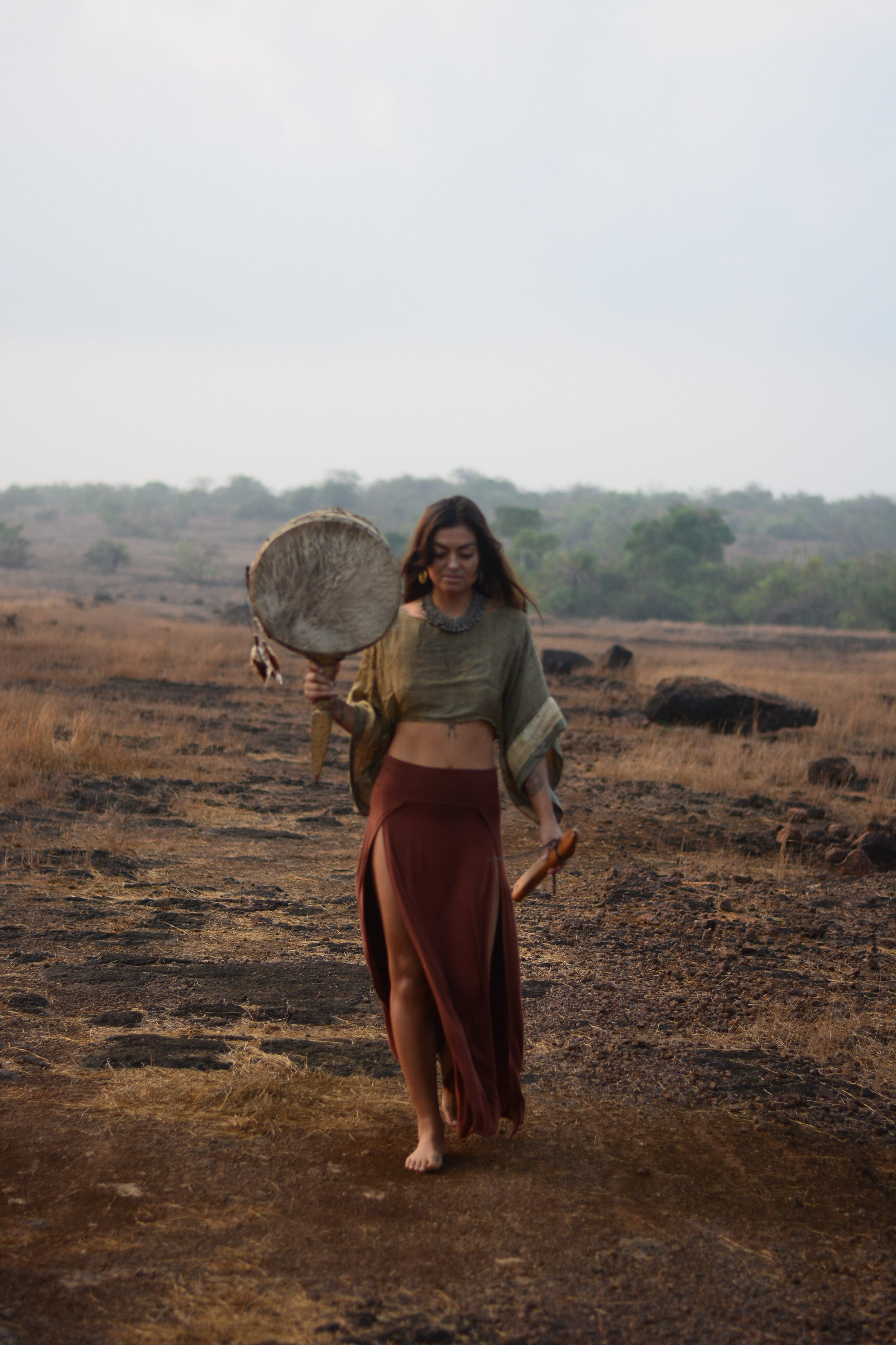 A woman walking barefoot in a dry, open field, carrying a large round tribal drum in her right hand and a smaller wooden percussion instrument in her left. She is wearing a crop top and a long skirt with high slits, and the landscape behind her featu
