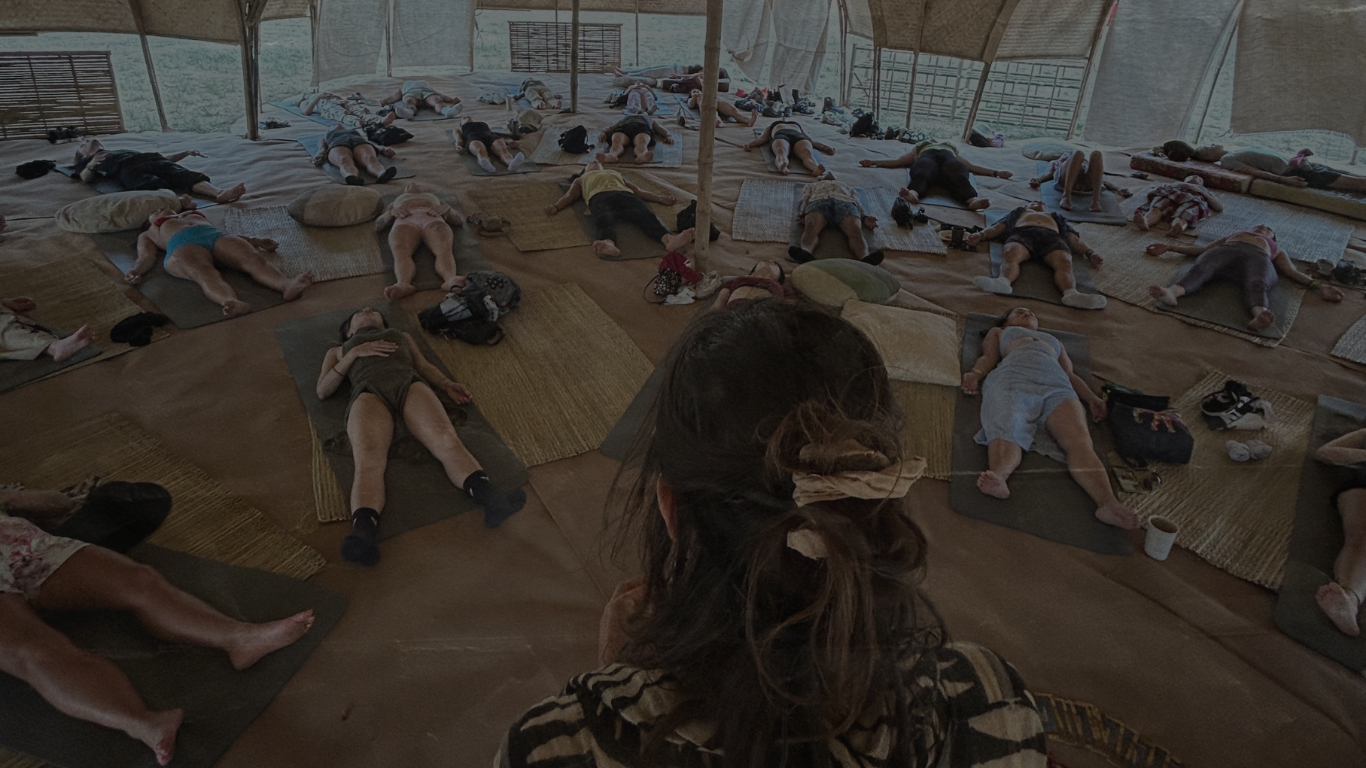 People lying on mats and pillows inside a spacious tent with bamboo or thatched roof, participating in a group meditation or yoga session.