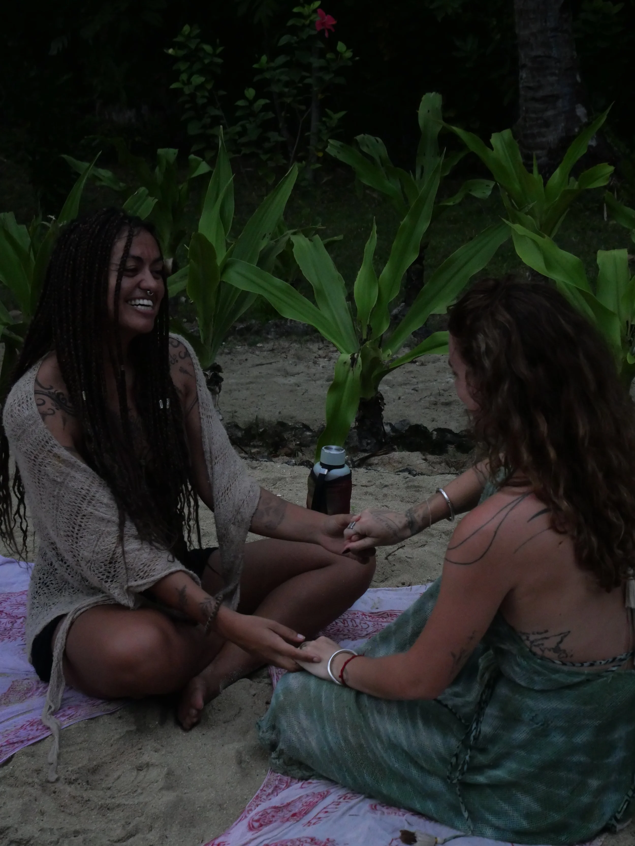 Kaylani with a woman sitting on beach towels by some corn plants, holding hands and smiling while engaging in a spiritual or ritual activity outdoors during an women's retreat.