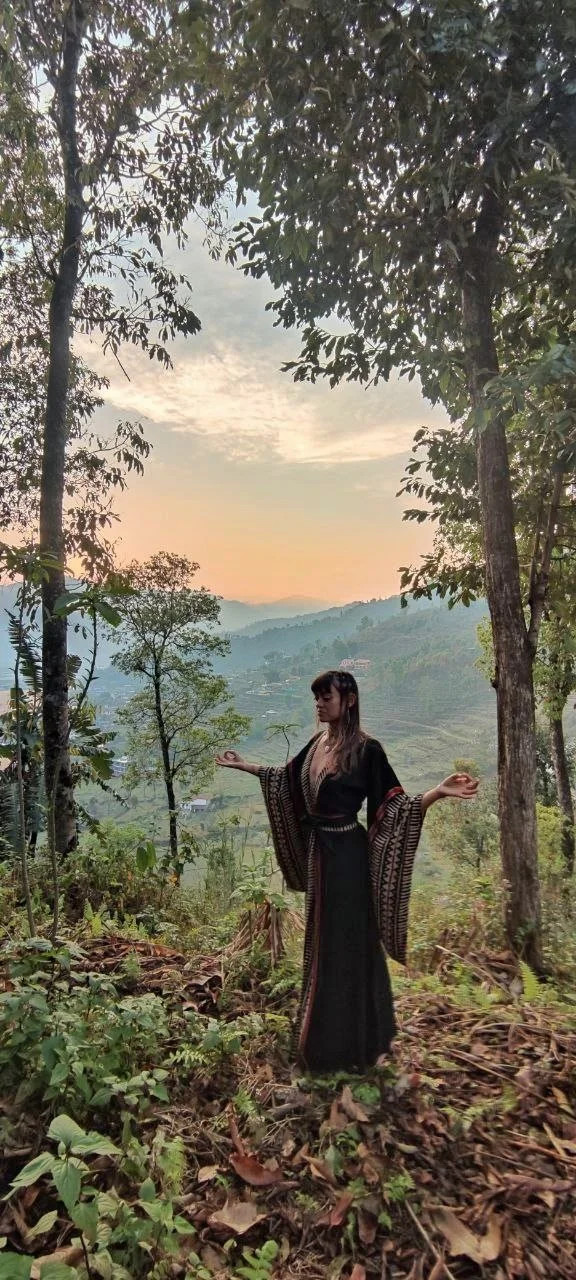 Kaylani in a black traditional dress standing in a forested area during sunset, with mountains and a village in the background. The Breath Temple.
