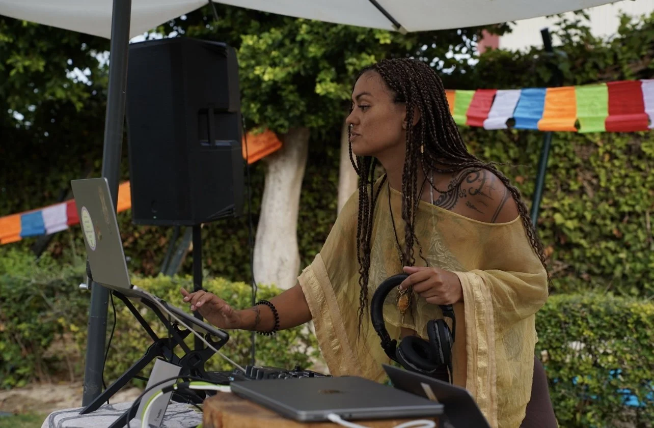 A woman (Kaylani) with braided hair and tattoos wearing a yellow top, using DJ equipment outdoors during a day-time ecstatic dance, with a large speaker, colorful flags, and trees in the background.