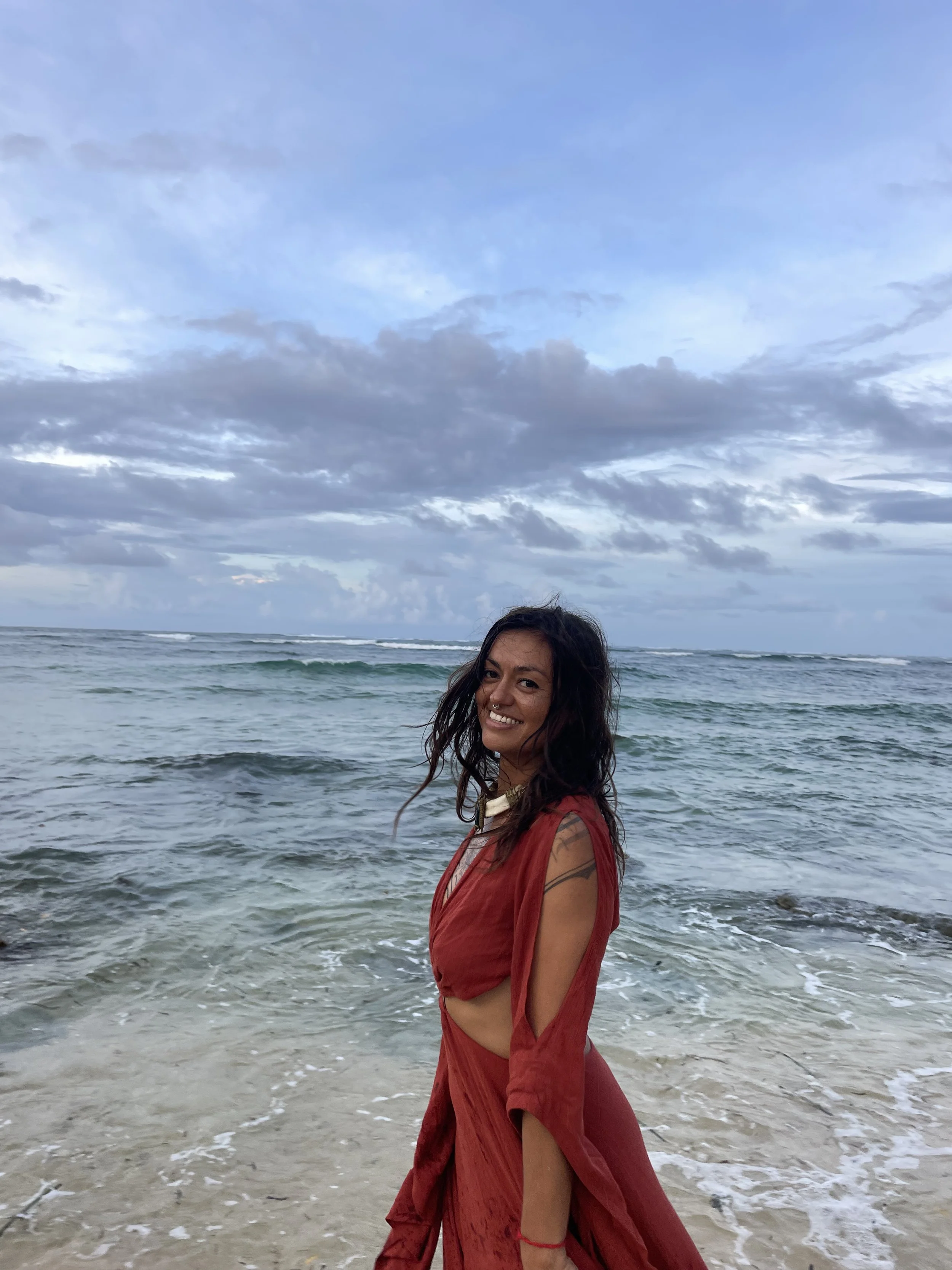 Woman, Kaylani, in a rust-colored dress smiling on the beach with ocean waves and cloudy sky in the background.