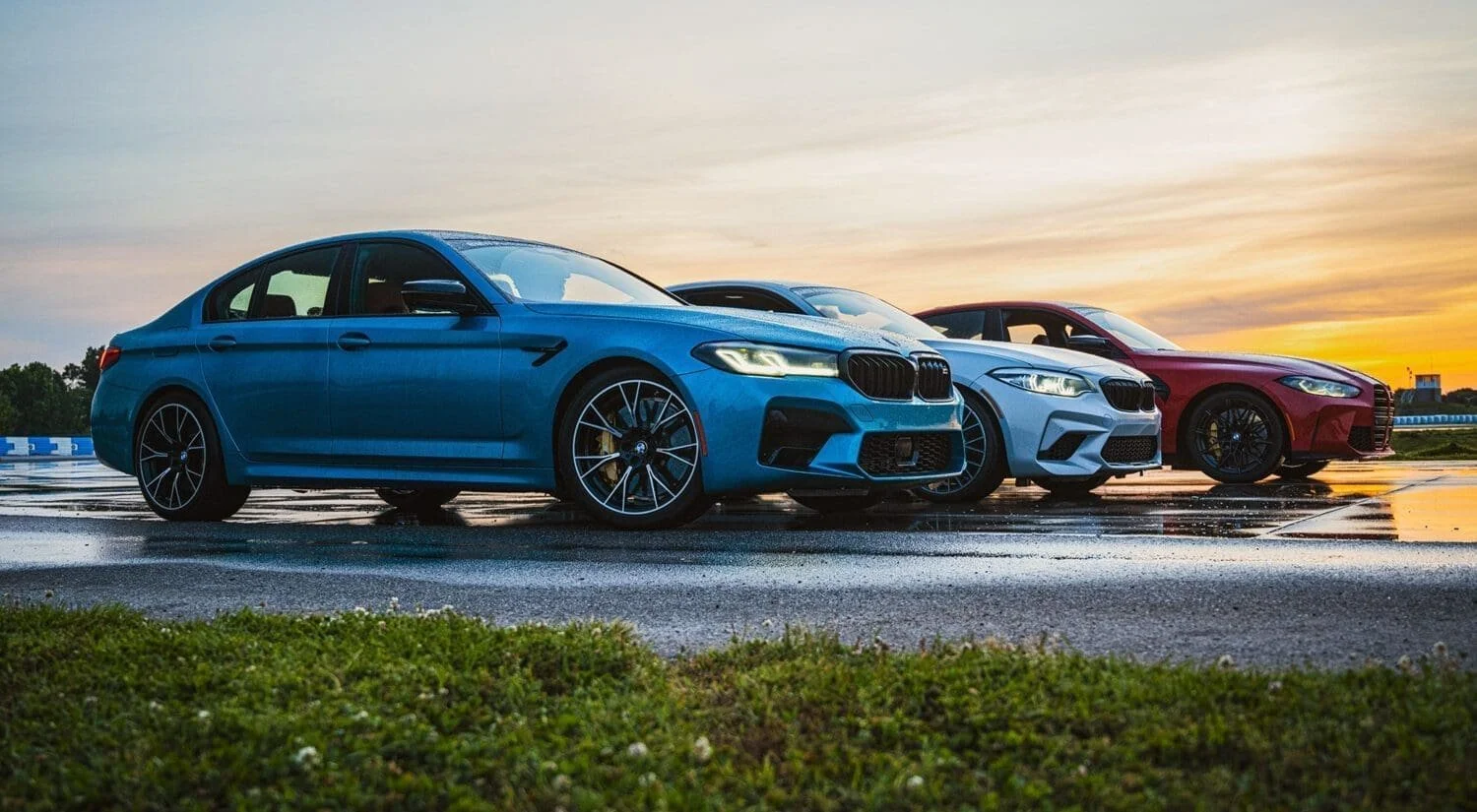 Three high-performance cars parked side by side on a wet race track during sunset, with grassy foreground and distant cityscape.