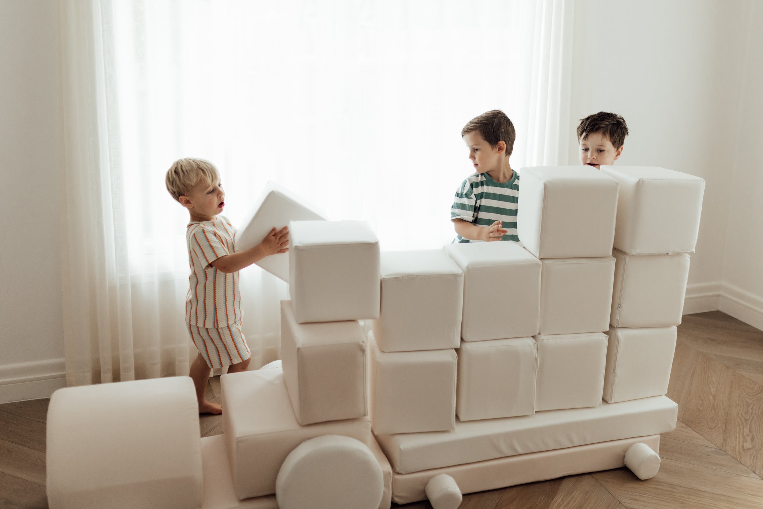 Three young children playing with a large white foam block structure in a bright room with hardwood floors and sheer curtains.