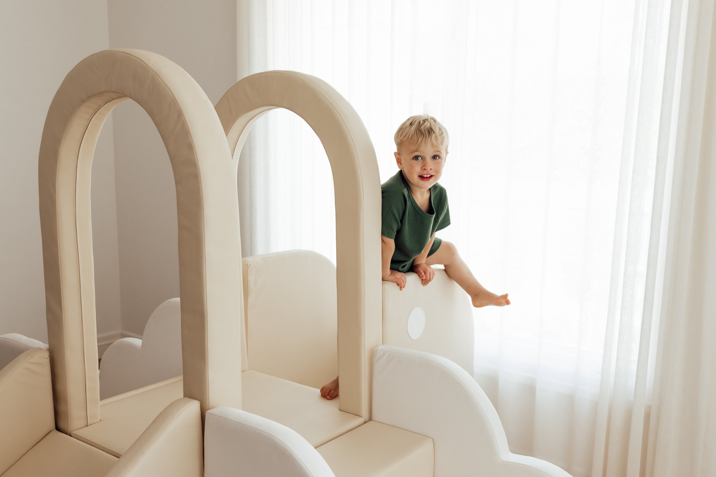 A young boy with blonde hair wearing a green shirt sitting on a white padded play structure near sheer white curtains.