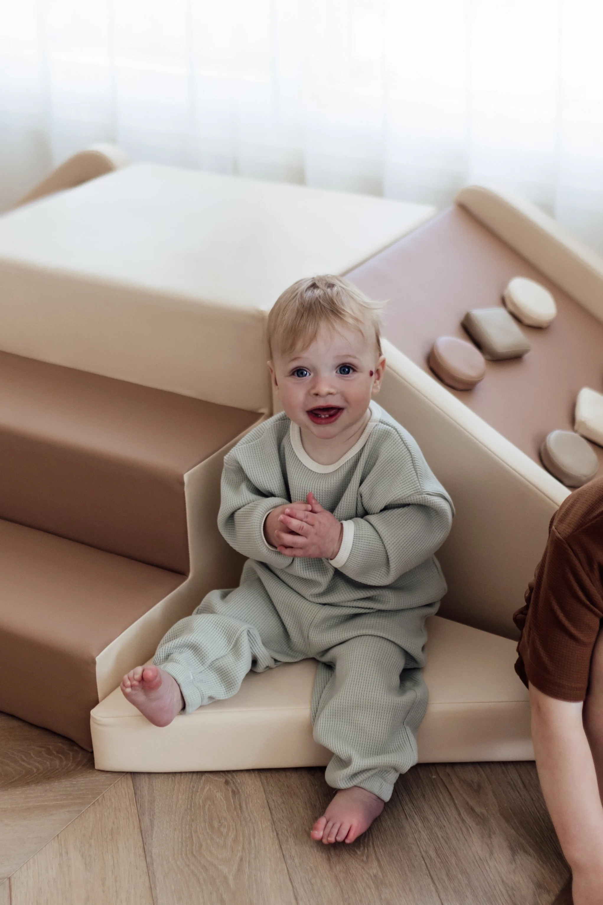 A young child with blue eyes and blonde hair sitting on a beige foam mat, smiling and clasping hands together in a living room with a wooden floor and a large window with sheer curtains in the background.