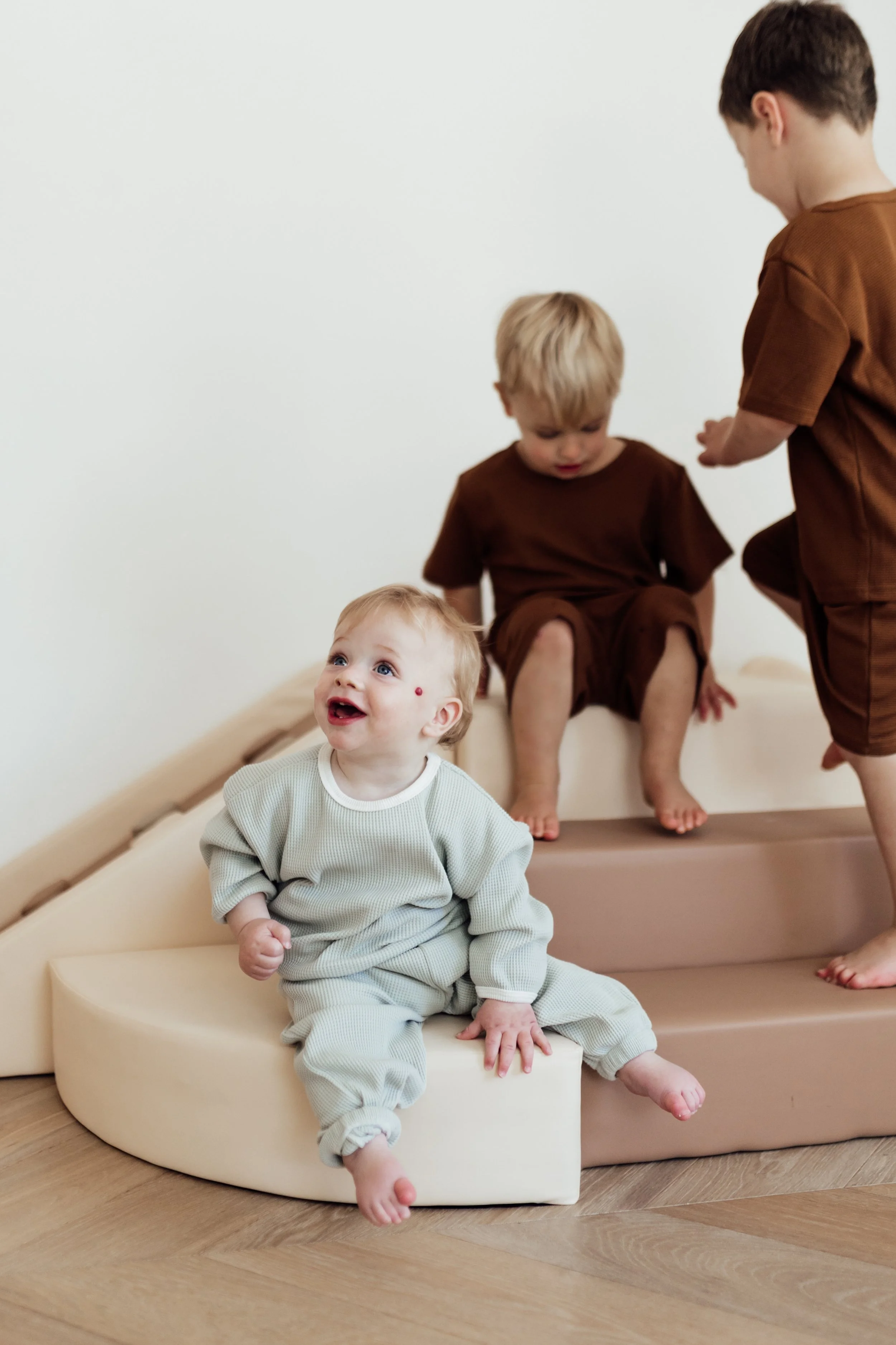 Three young children playing on soft steps inside a room with wooden flooring and white walls.