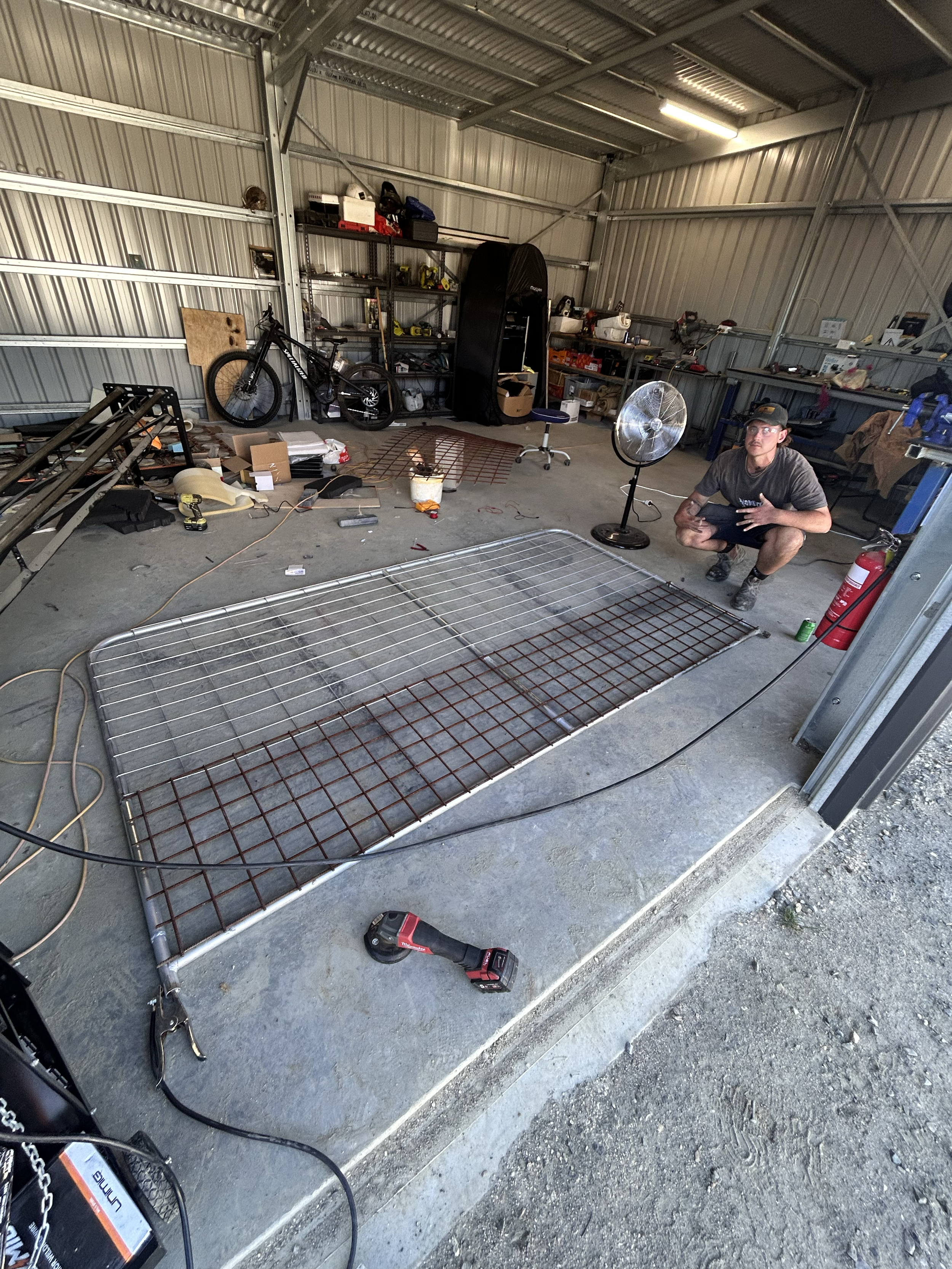 A garage with a man crouching near a fire extinguisher, a fan, and a partially assembled custom fence frame on the floor. Shelves behind him hold tools, equipment, and a bicycle. The garage has a concrete floor and metal walls.