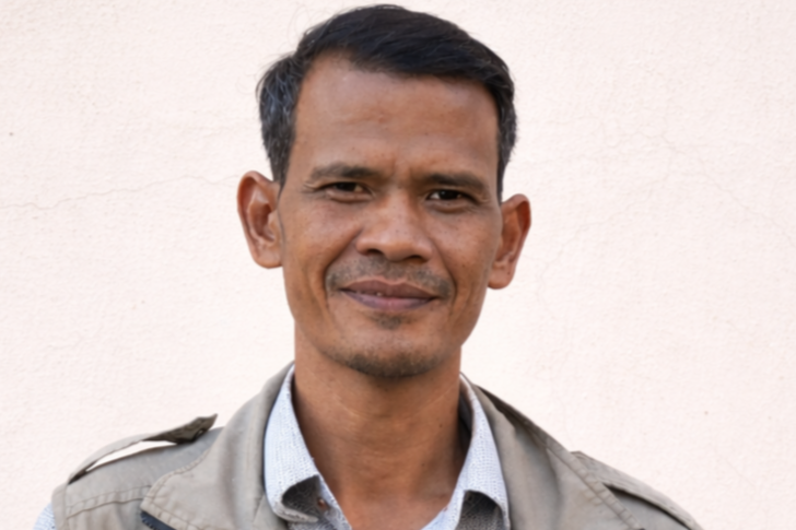 A middle-aged man with short dark hair, wearing a light gray jacket over a collared shirt, standing outdoors in front of a plain wall.