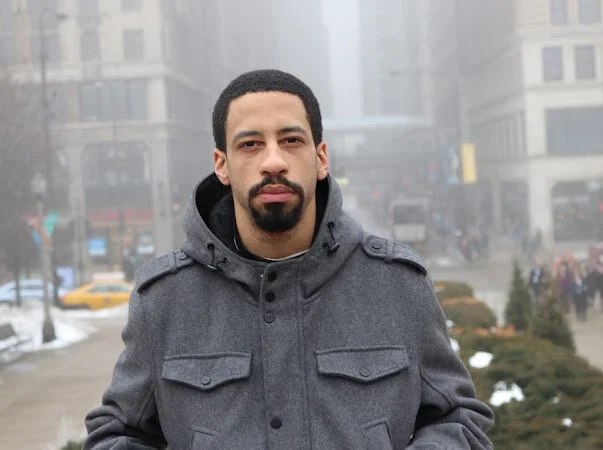 Young man with a beard and mustache wearing a gray jacket and hood, standing outdoors in an urban area with buildings and fog in the background.