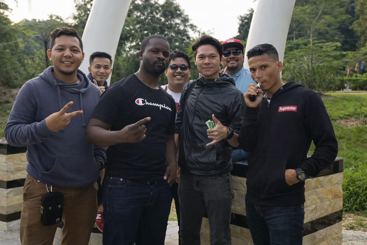 A group of seven young men standing outdoors, some making peace signs and holding objects, with greenery and large white structure in background.
