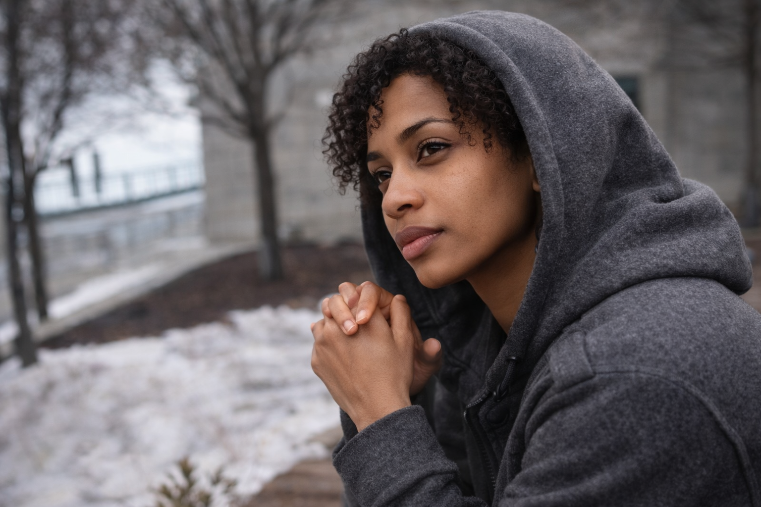 A woman in a gray hoodie with hands clasped, sitting outdoors on a cold day with snow on the ground and trees in the background.