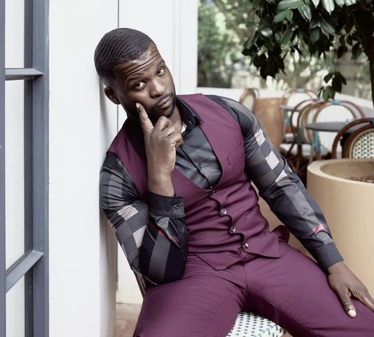 A man in a maroon vest and checkered shirt sitting on a chair outdoors, posing with his hand on his chin and looking at the camera.
