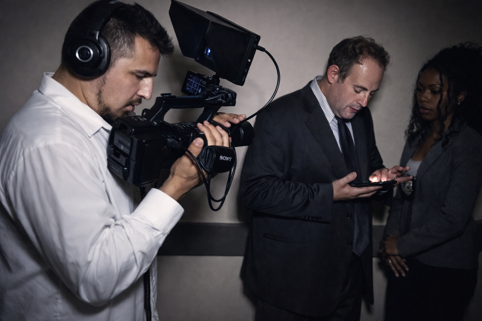 A cameraman filming a scene with two people in a dark setting, one man using a phone and a woman standing nearby.