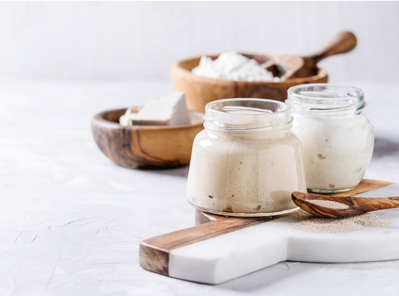 Two glass jars of cream or yogurt, wooden bowls with powder and a block of white cheese, and wooden spoons on a white textured surface.