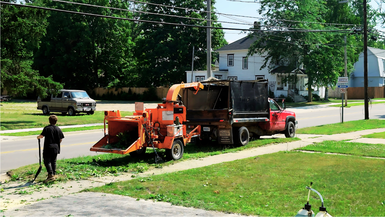 A West Michigan Tree Services ground crew member clears debris near a wood chipper and truck, illustrating the organized 'Ground Support System' necessary for safe and efficient residential tree removal.