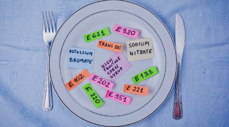 A white dinner plate covered in colorful sticky notes labeled with common food additives like Potassium Bromate, High Fructose Corn Syrup, and Sodium Nitrate, visualizing hidden ingredients in the modern food supply.