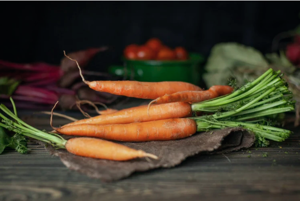 A bundle of fresh, vibrant orange organic carrots with leafy green tops resting on a rustic wooden table, representing the nutrient density and soil health benefits of choosing organic produce over chemically treated alternatives.