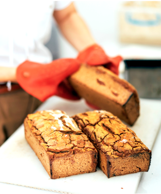 Two slices of marble cake and one loaf of marble cake on a white surface, with a person in the background using a red oven mitt to hold a baking pan with the cake.
