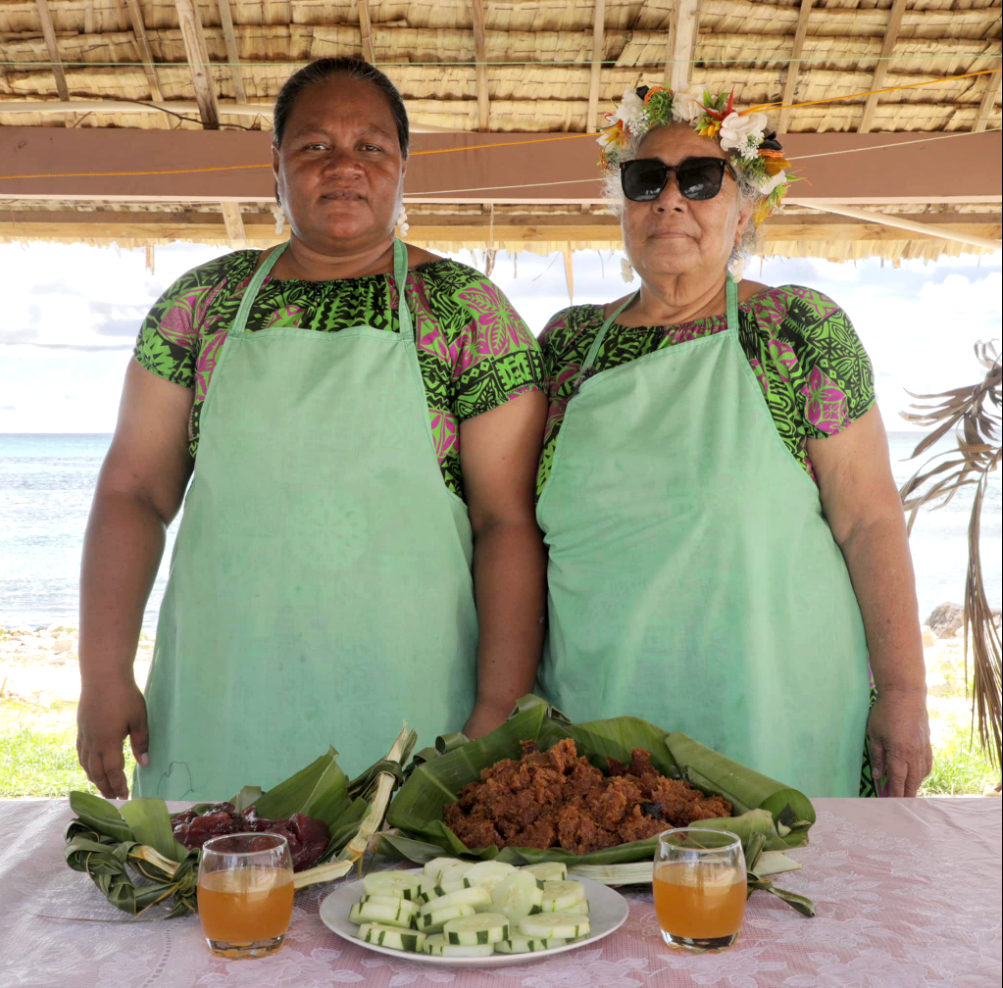 Two women from Nukulaelae and Funafuti, Tuvalu, wearing traditional floral garlands and aprons, standing behind a heritage meal of taro pudding and fresh coconut toddy.