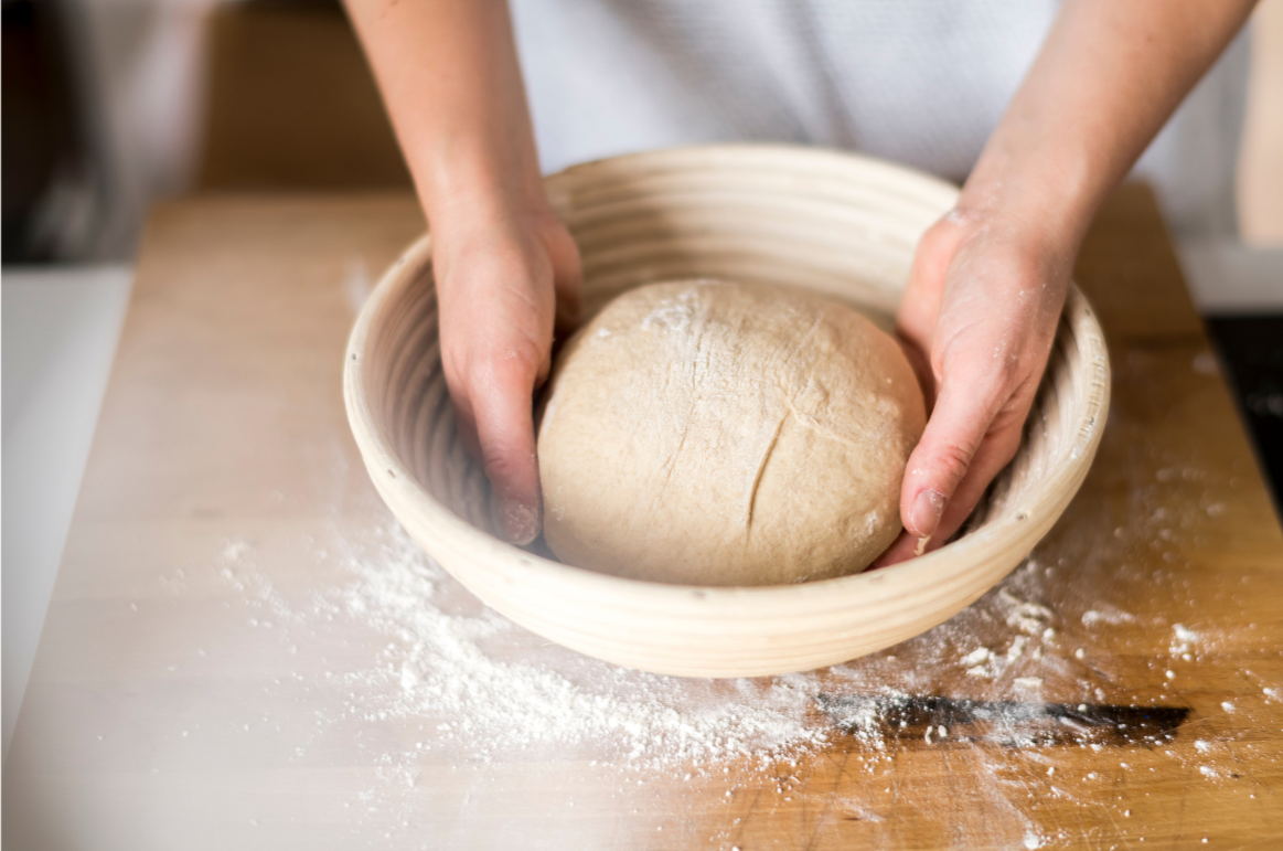 Person shaping dough in a wooden bowl, with flour scattered on a wooden countertop.