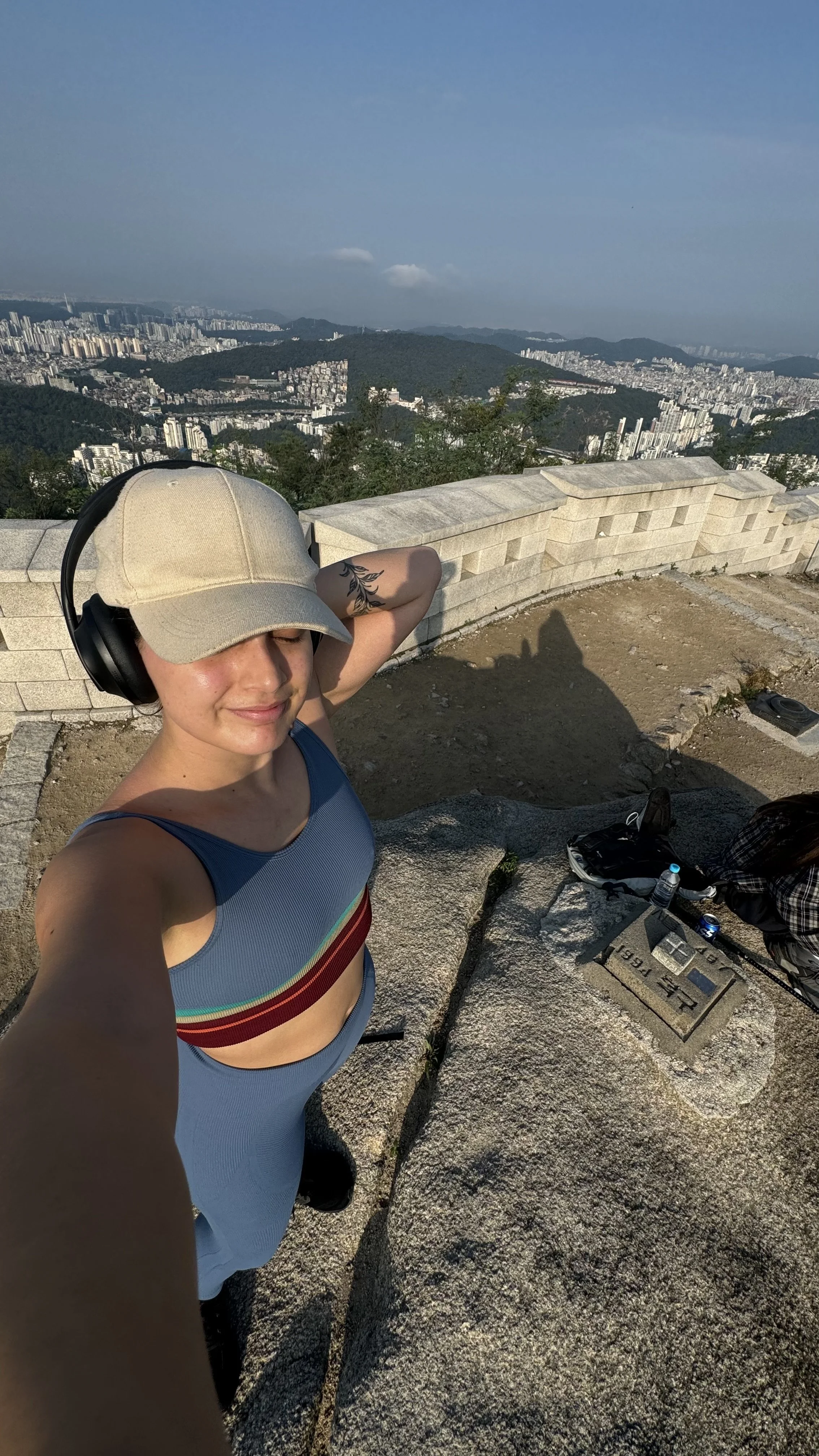 Selfie de uma mulher com boné, fone de ouvido e roupa de ginástica, com uma vista de cidade e montanhas ao fundo.