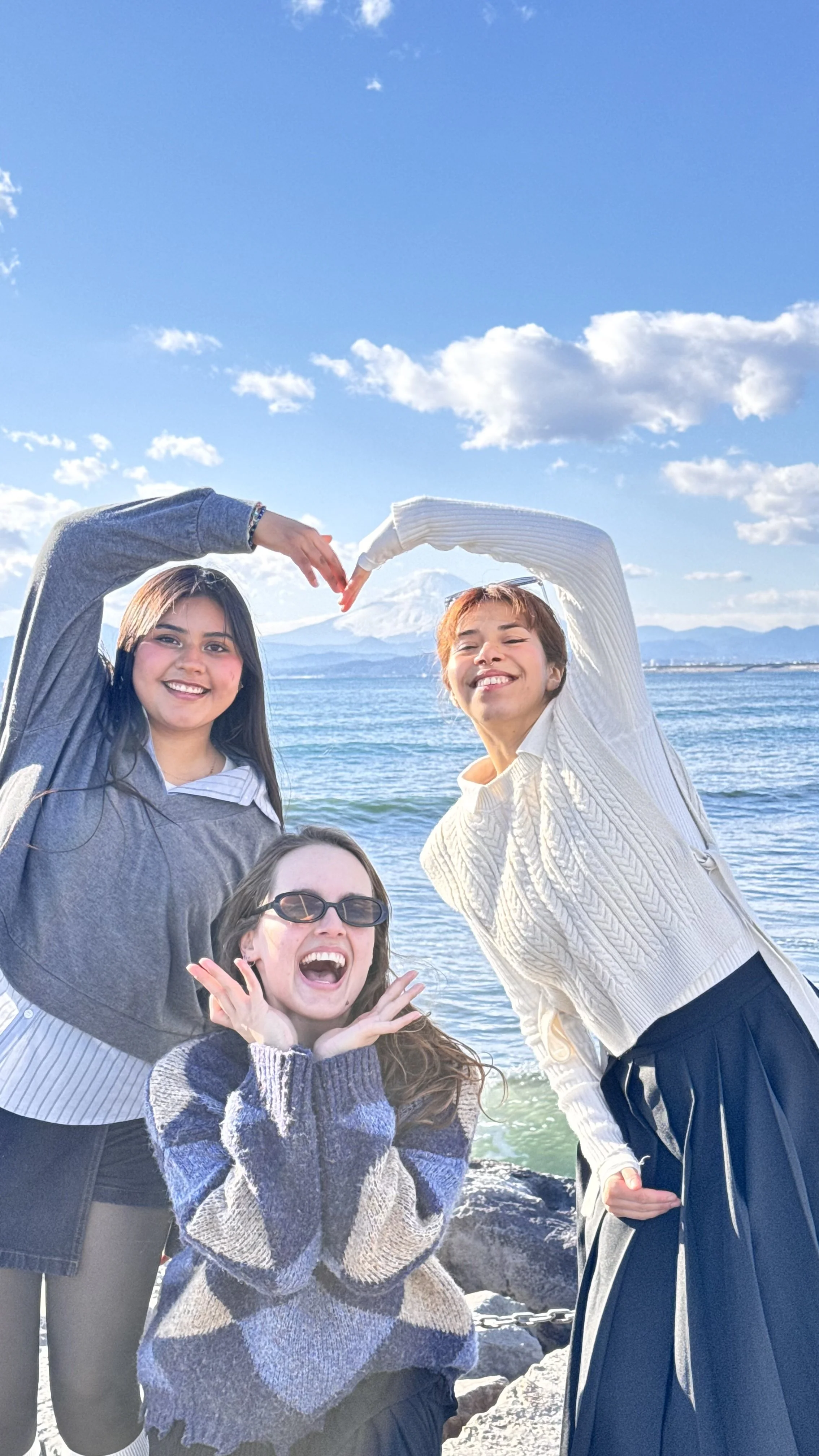 Três jovens sorrindo na praia, com uma mulher na frente, duas pessoas formando um coração com as mãos ao fundo, vista para o mar e montanhas ao longe.