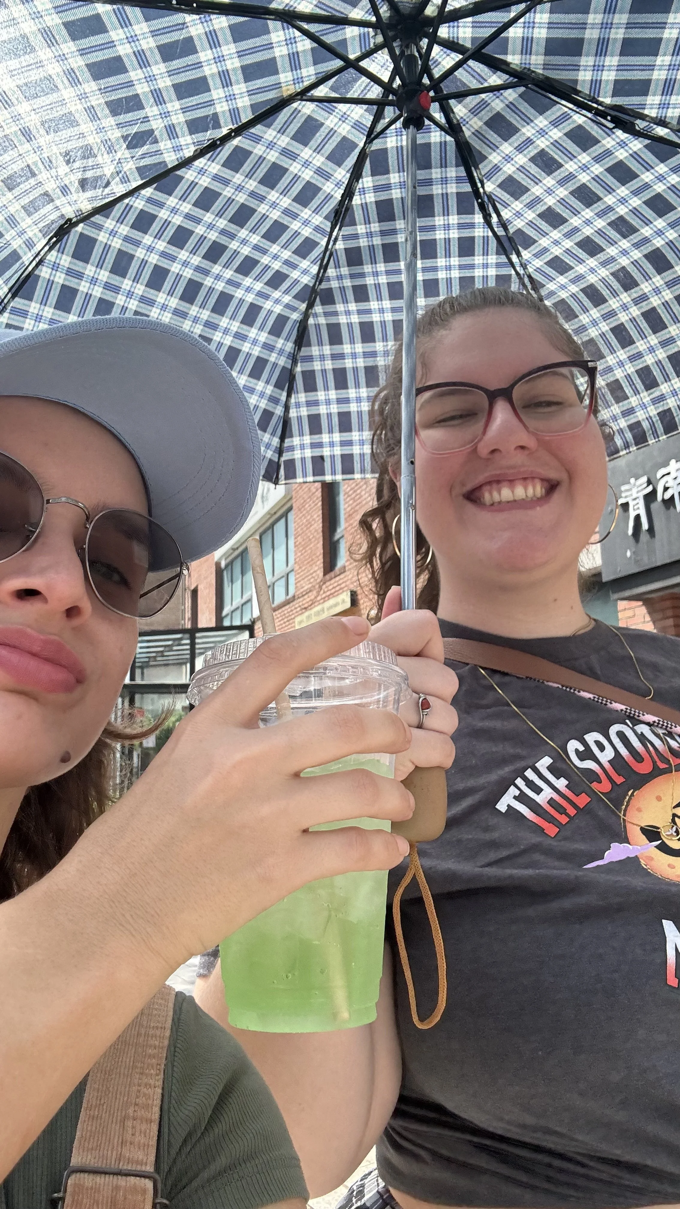 Duas mulheres sorridentes sob um guarda-chuva de quadriculados, uma delas segurando uma bebida verde em copo plástico com tampa e canudo, em ambiente urbano ao ar livre.