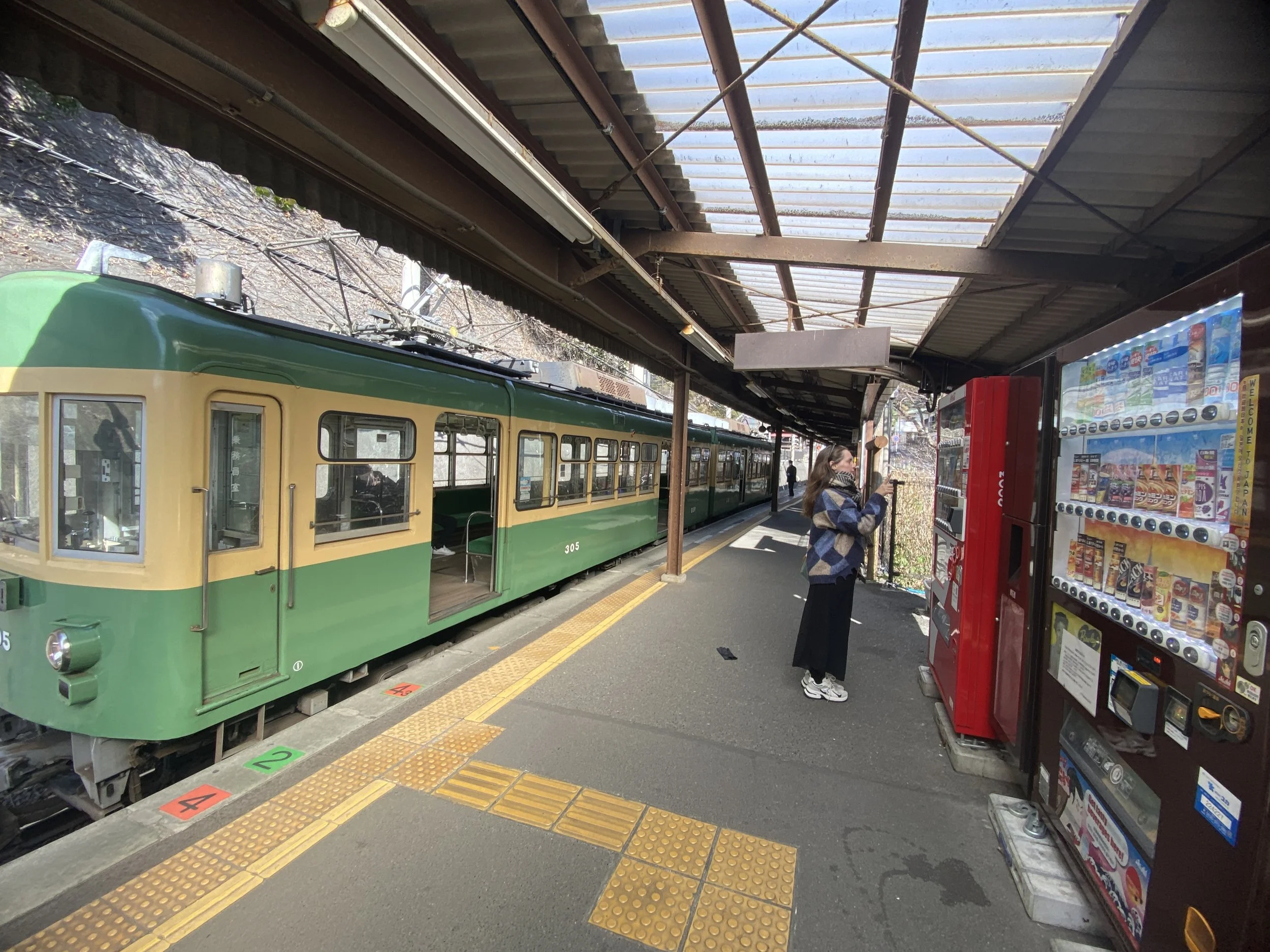Pessoa usando máscara comprando junto a uma máquina de venda automática na plataforma de um bonde antigo, ao lado de um trem verde e amarelo. A plataforma possui piso tátil yellow e marked com cores vermelha, verde, amarela e preta.