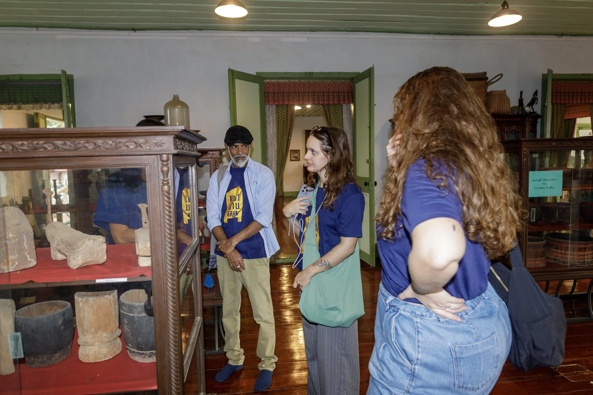 Quatro pessoas conversando dentro de uma sala de museu com exibição de antiguidades, algumas exibidas em uma vitrine de madeira. As pessoas estão vestidas com camisetas azuis e uma delas segura um microfone.