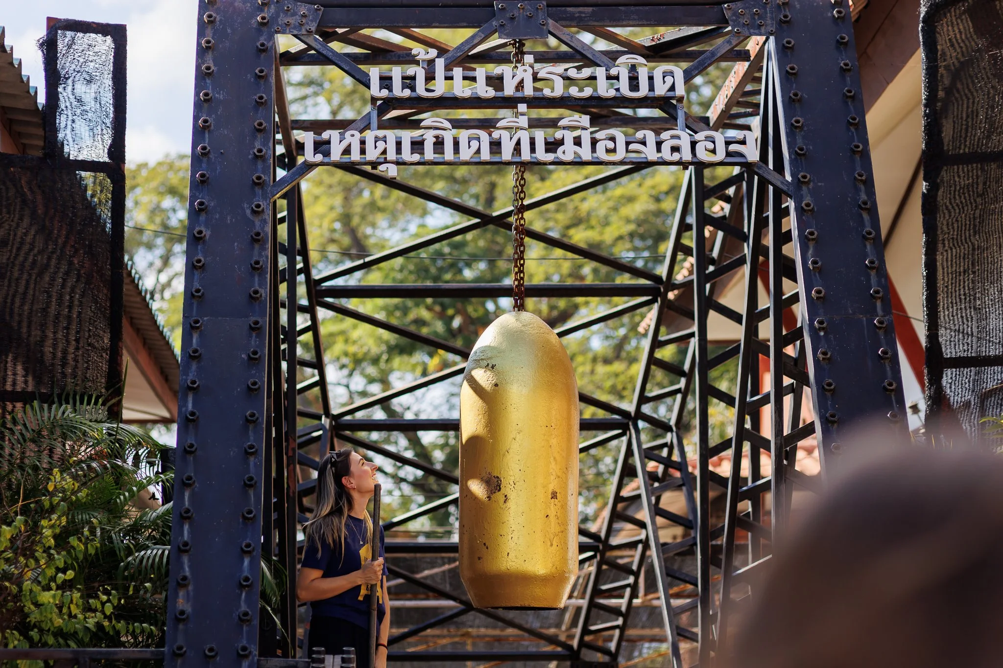 Pessoa observando uma estrutura de ferro com uma grande cápsula dourada pendurada no centro, ao ar livre, com árvores ao fundo e uma placa com escrita em tailandês.
